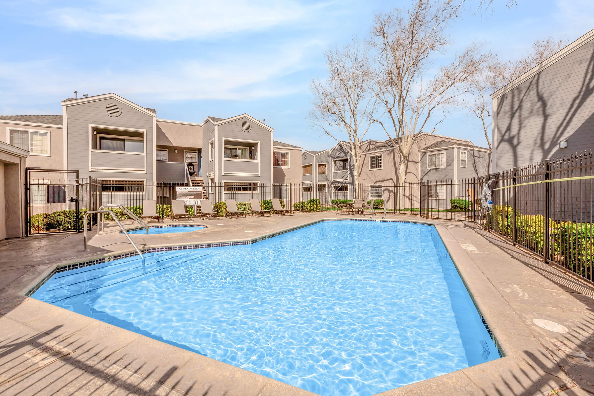 A swimming pool surrounded by a fenced area, with lounge chairs and potted plants. Nearby buildings in a residential complex are visible, featuring multiple stories and balconies under a clear blue sky. Trees with bare branches are in the background.