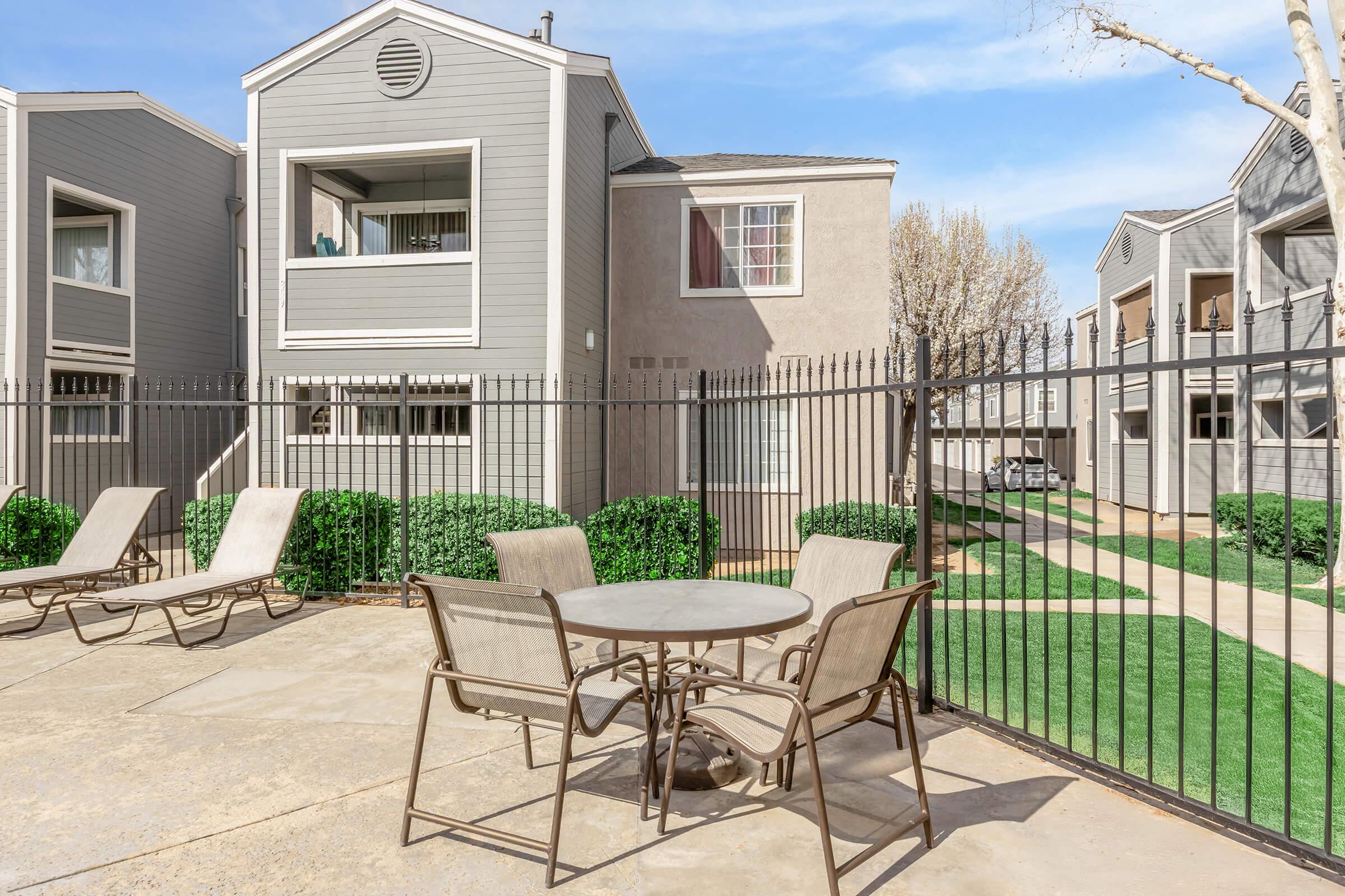 A view of a residential apartment complex featuring two buildings with balconies, surrounded by a black metal fence. A small patio area includes a round table and four chairs, with lounge chairs nearby, set against a backdrop of well-maintained greenery and a clear blue sky.