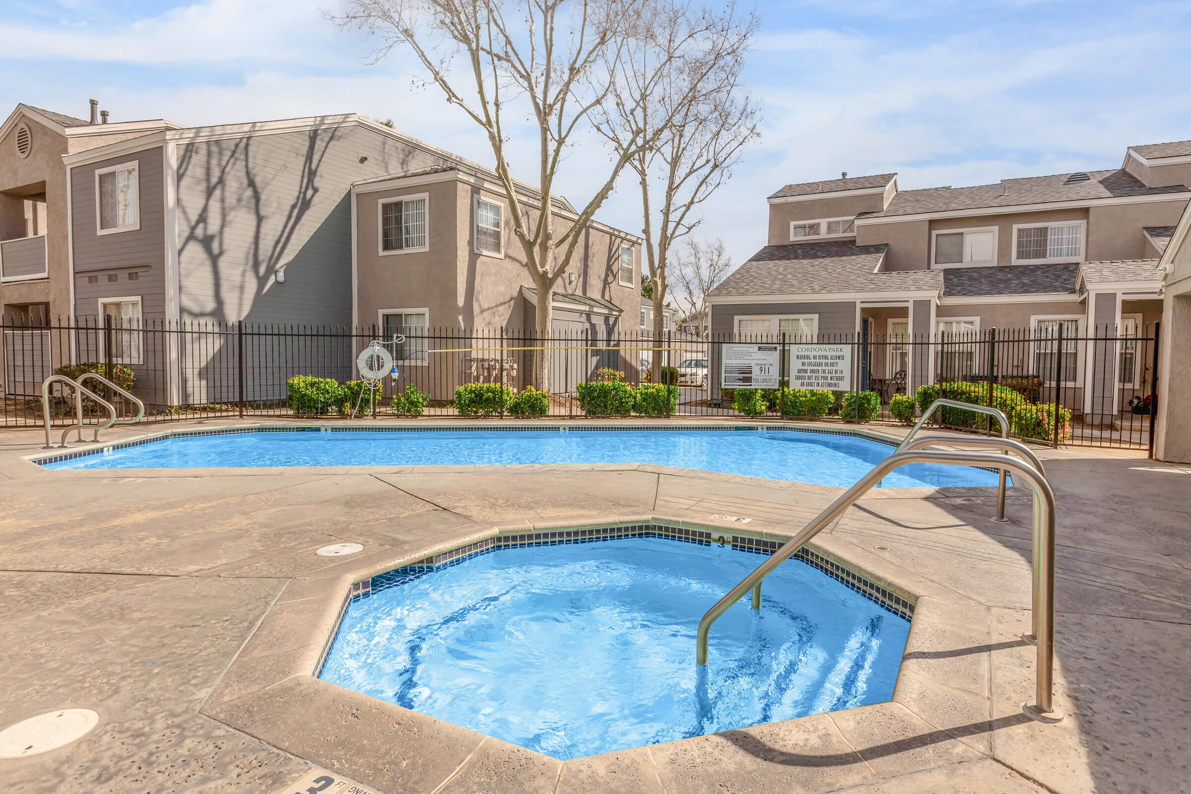 A sunny view of a residential swimming pool area featuring a large rectangular pool and a smaller octagonal hot tub. Surrounding the pool are manicured bushes and trees, with a fence in the background. The scene conveys a relaxing outdoor atmosphere in a community setting.