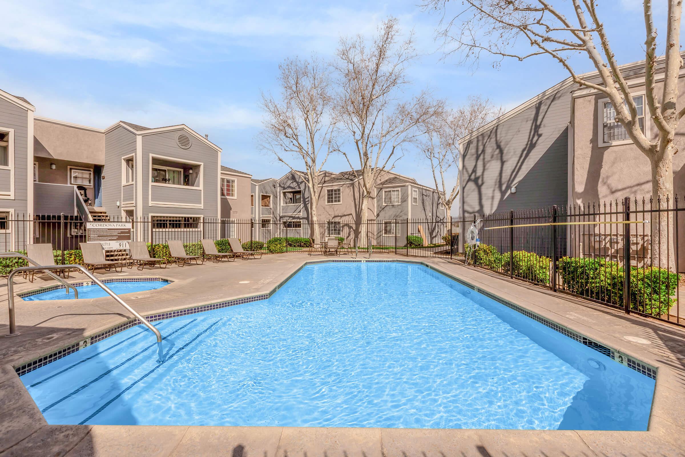 A clear, inviting swimming pool surrounded by sun loungers and fenced by low greenery, with residential buildings in the background. The scene is well-lit and showcases trees with sparse foliage, suggesting a pleasant outdoor setting suitable for relaxation.