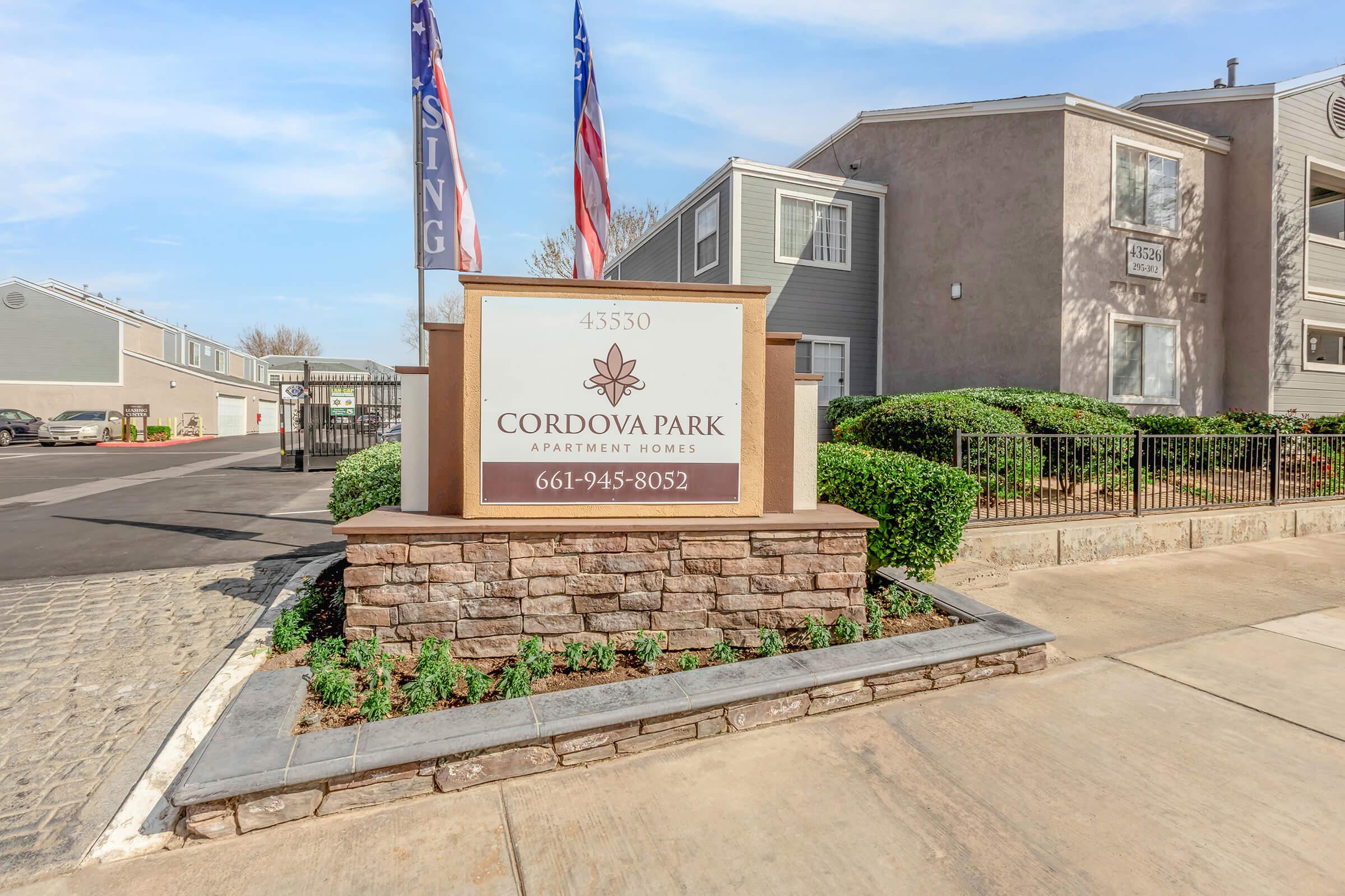 Sign for Cordova Park Apartment Homes, located at 45350. The sign features a decorative design, surrounded by trimmed bushes, with flags in the background. The apartment complex is visible, along with a paved pathway and parking area.
