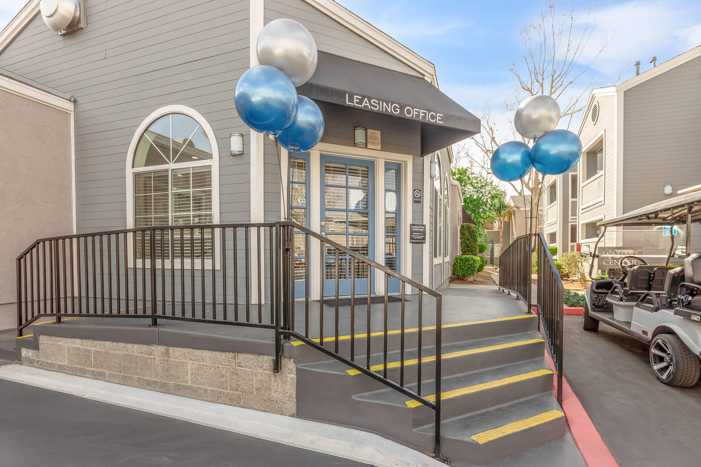 A leasing office entrance featuring a ramp and decorative balloons in blue and silver. The building has large windows and a sign indicating it's a leasing office. A golf cart is parked nearby, and there is landscaping visible in the background. The sky is clear and bright, suggesting a pleasant day.