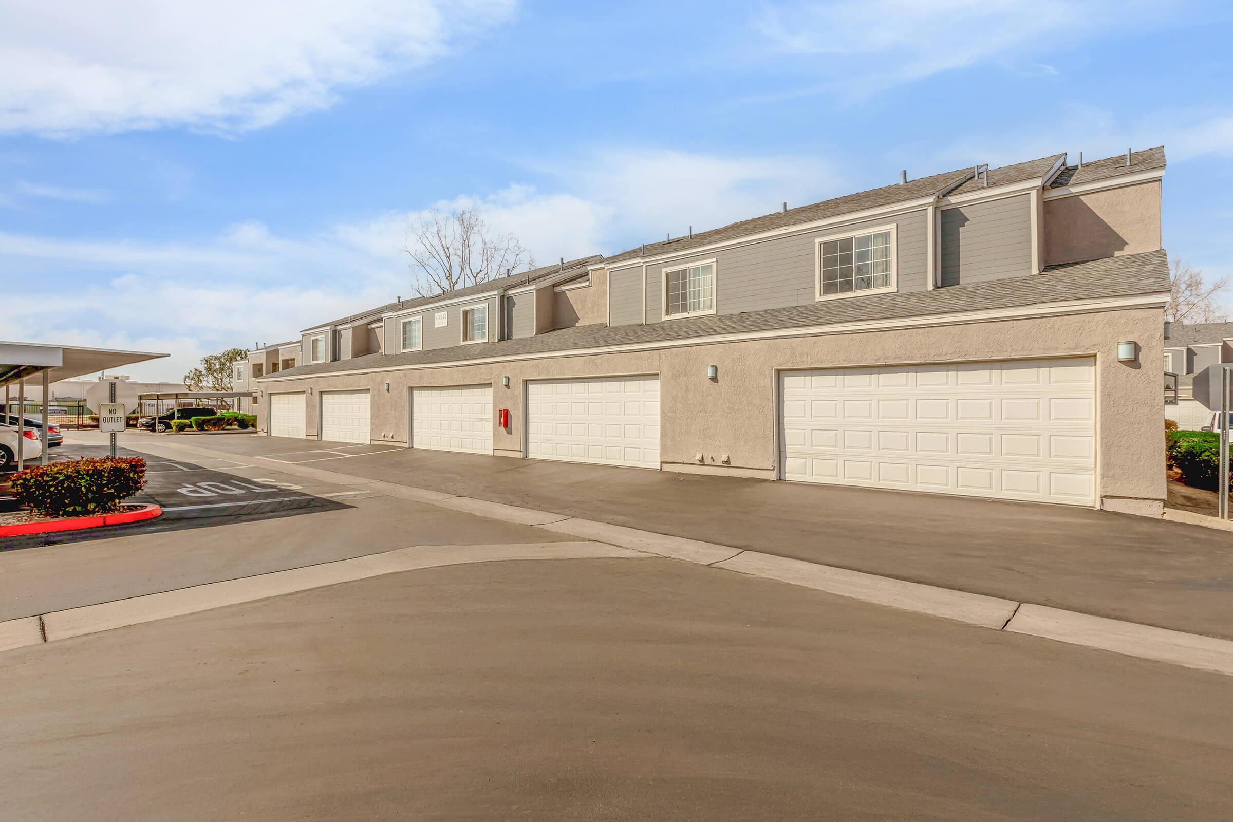 A row of residential buildings with attached garages, situated in a well-kept area. The pavement is smooth, and there are bushes along the edge. A parking area is visible nearby, and the sky is partly cloudy, creating a serene atmosphere.