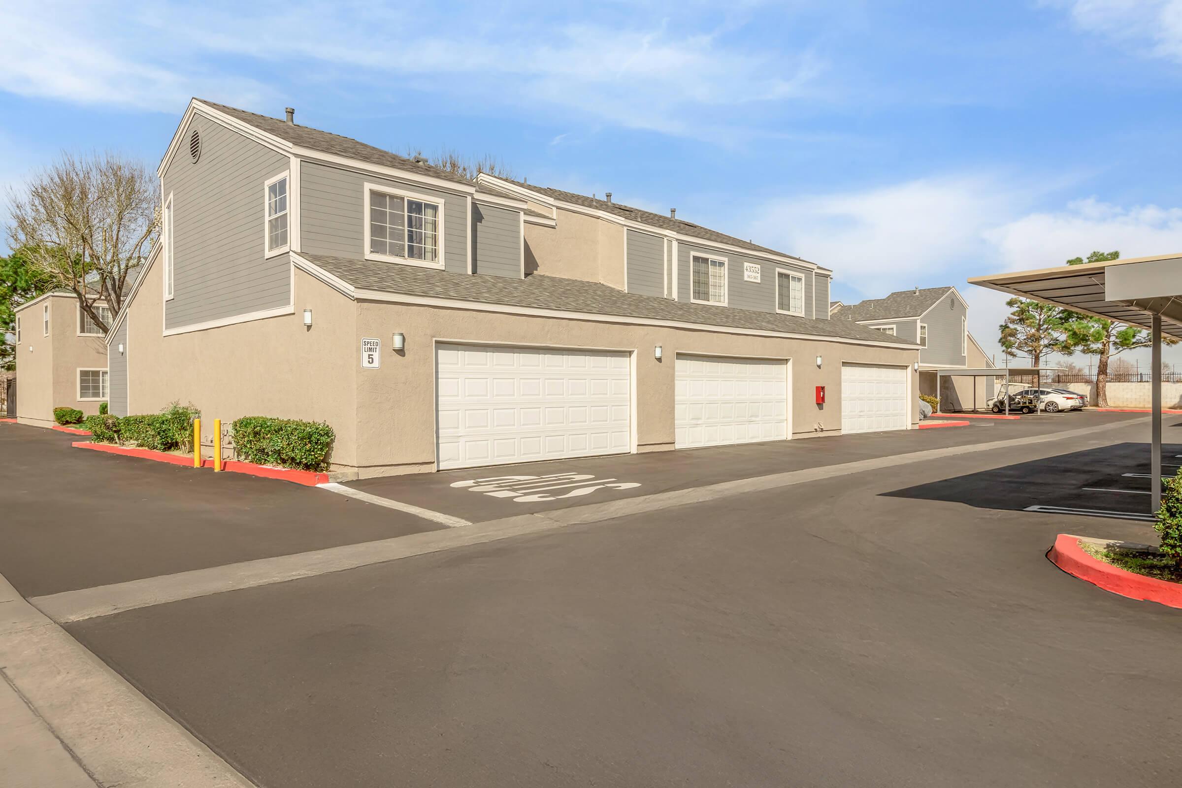 A view of a residential community with two-story apartment buildings featuring gray exteriors. There are garages on the ground level, well-maintained landscaping along the entrance, and a clear blue sky above. The driveway is paved, and there are designated parking spots visible in the background.