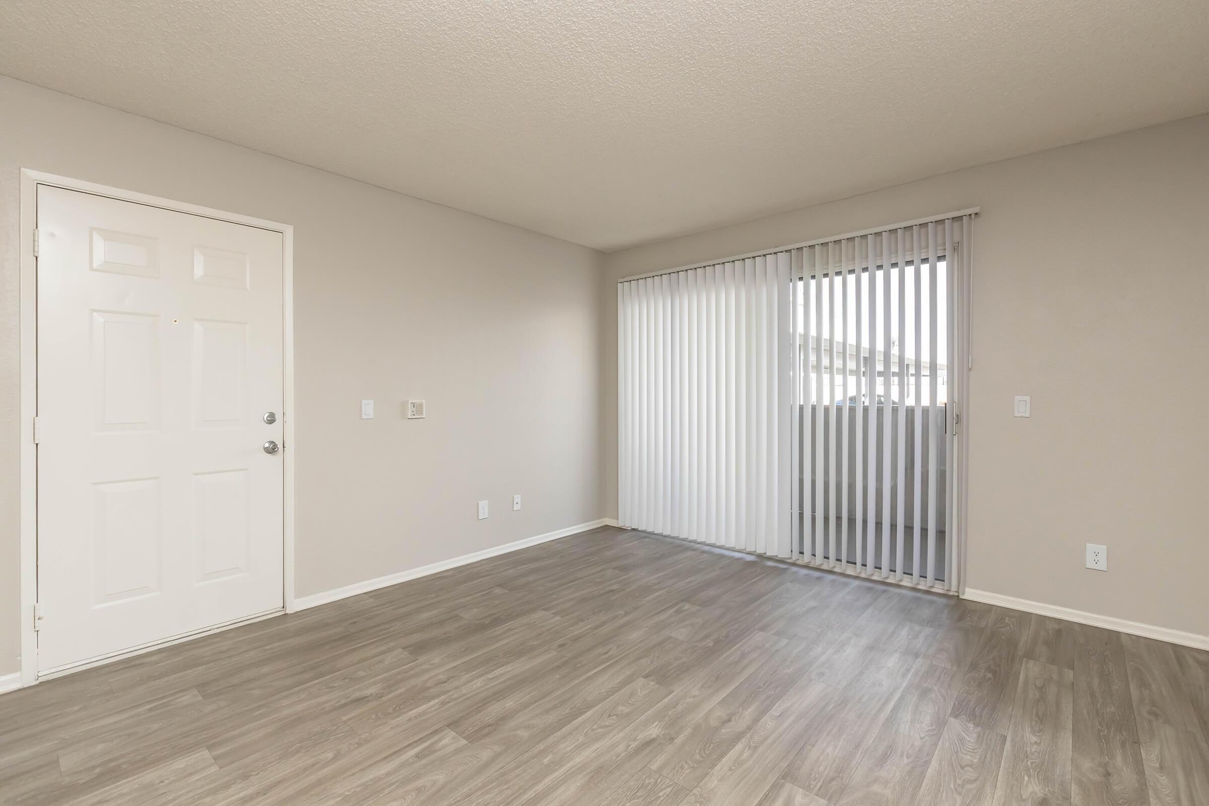 A spacious, unfurnished living room featuring light-colored walls, a front door on the left, and large sliding glass doors with vertical blinds leading to a balcony. The floor is covered in light brown laminate, providing a clean and neutral ambiance.