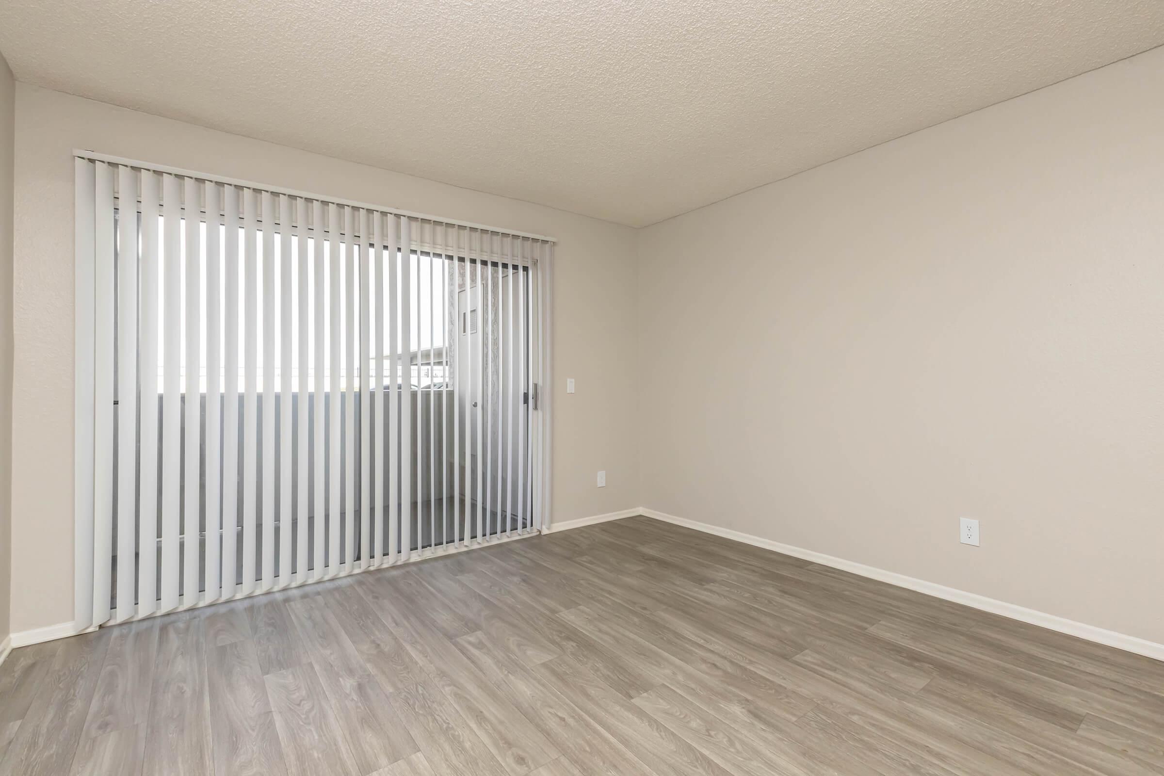 Empty room with light-colored walls and a large sliding door covered with vertical blinds, leading to a balcony. The floor is a light laminate wood, and the space is bright with natural light coming through the door. There are no furniture or decorations, creating an open and spacious feel.