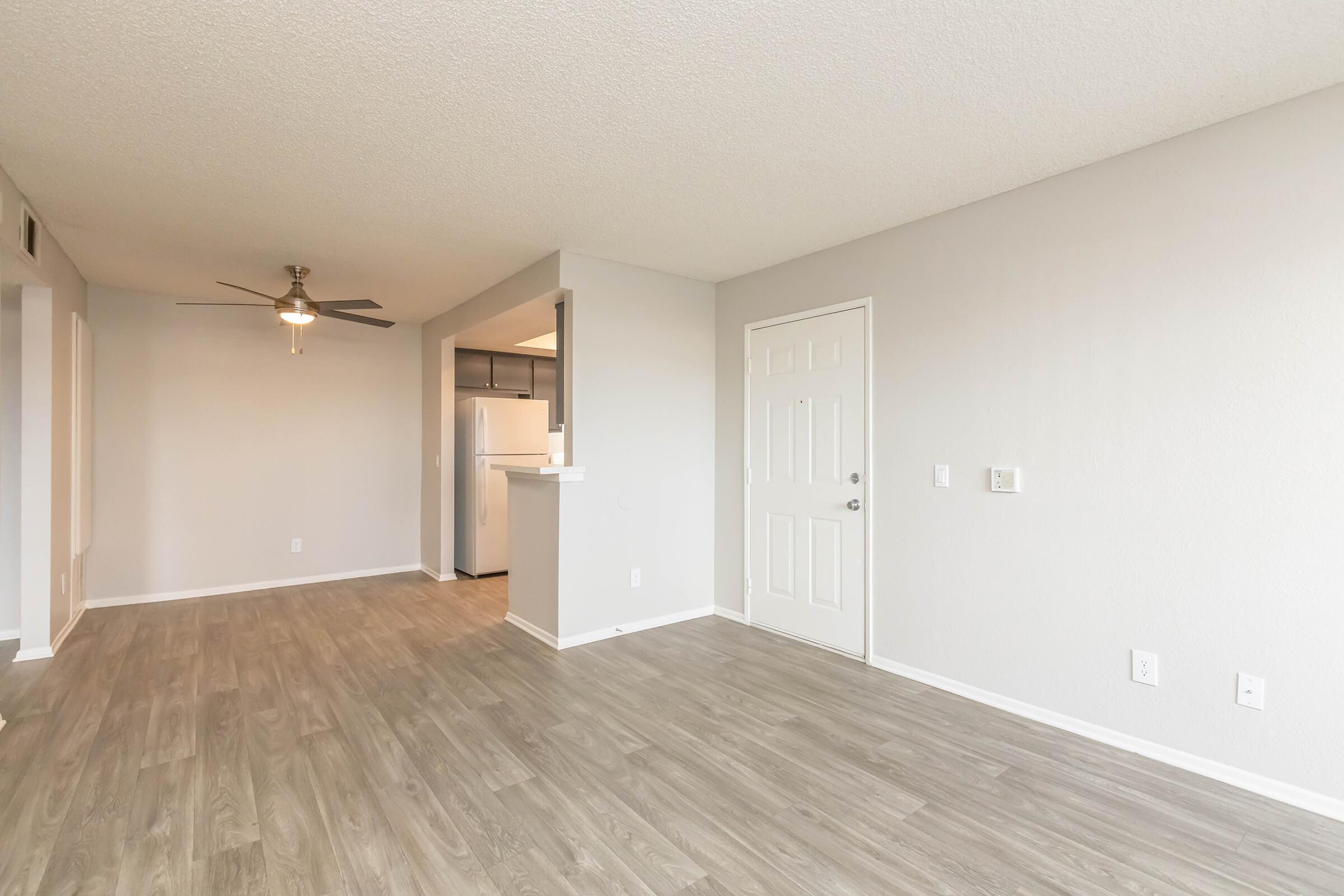 Spacious living area with light beige walls and wood-like laminate flooring. Features a ceiling fan and a doorway leading to a kitchen with white appliances. The entrance door is visible, and the overall ambiance is bright and inviting.