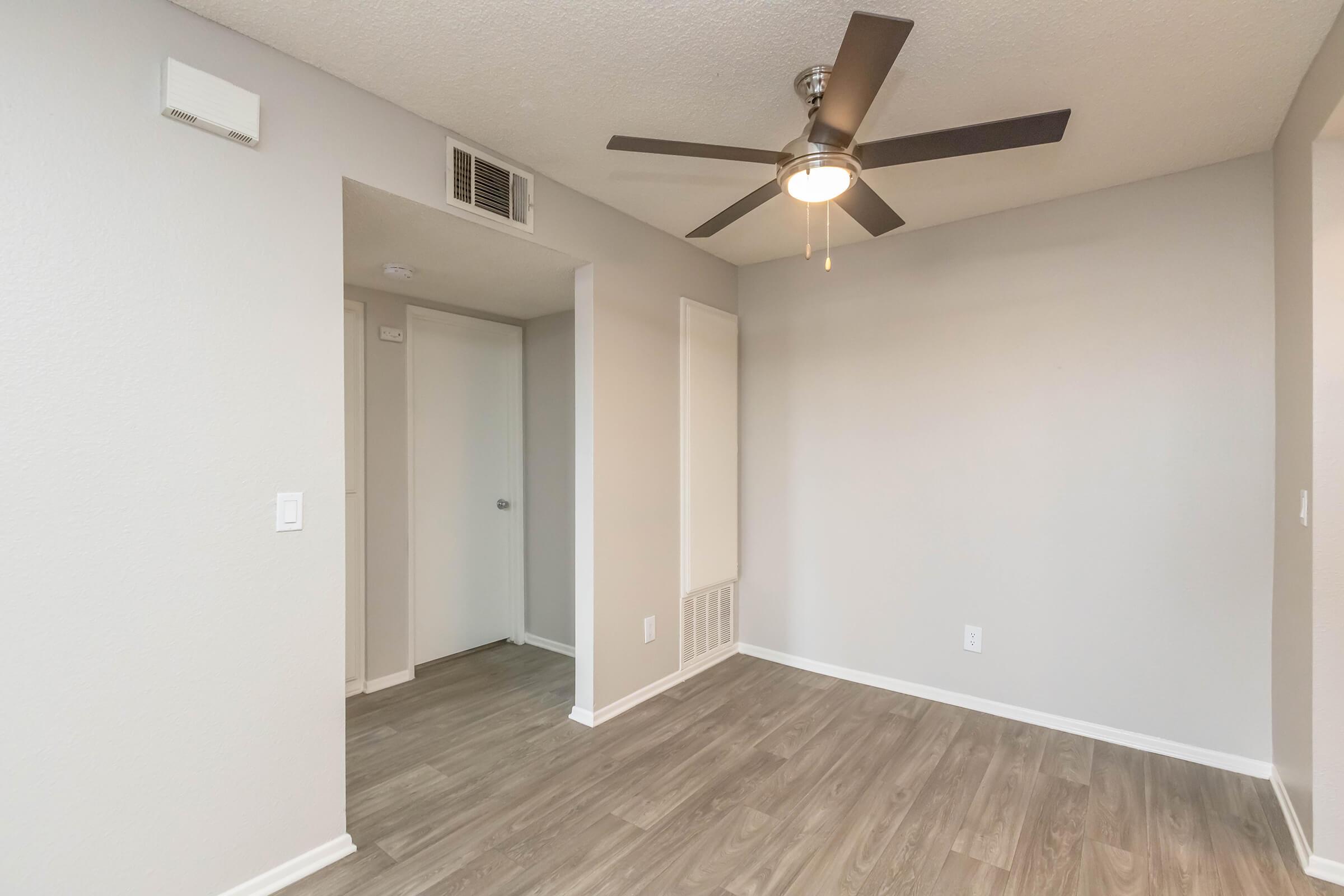 A spacious, light-filled room featuring a ceiling fan, neutral gray walls, and wood-like flooring. There is a door leading to another room and a small wall vent. The overall atmosphere is modern and inviting, suitable for a living or dining area.