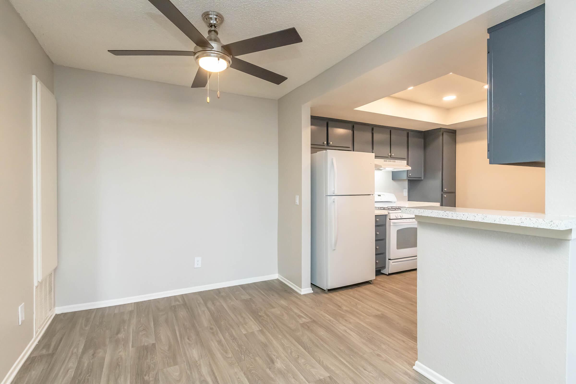 Interior of an apartment featuring a light gray wall, a ceiling fan, and a kitchen area with white appliances, including a refrigerator and stove. The flooring is a warm wood-like material, and the adjacent wall has dark blue cabinets. Natural light enters the space, creating a bright and inviting atmosphere.