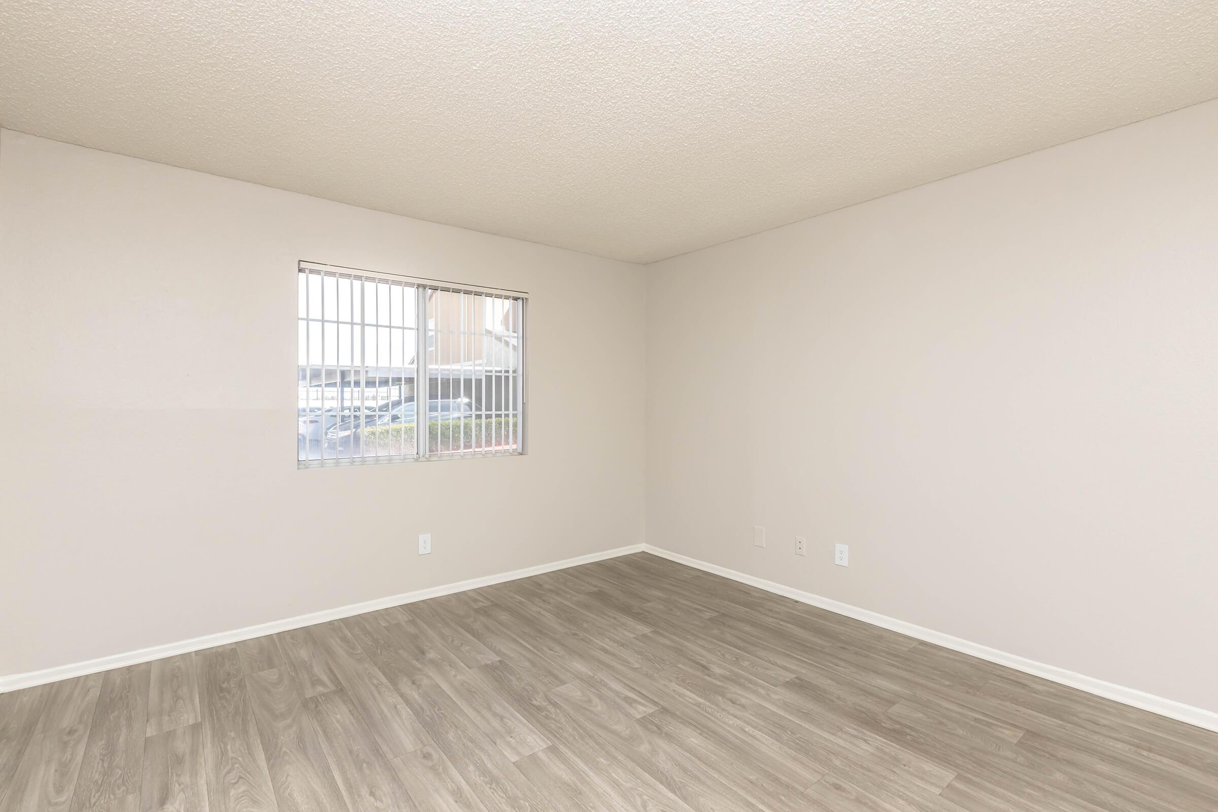 Empty room with light-colored walls and a window featuring vertical blinds. The floor is covered in light wood-like laminate. There are no furnishings or decorations, creating a spacious and minimalistic feel. Natural light enters through the window, illuminating the interior.