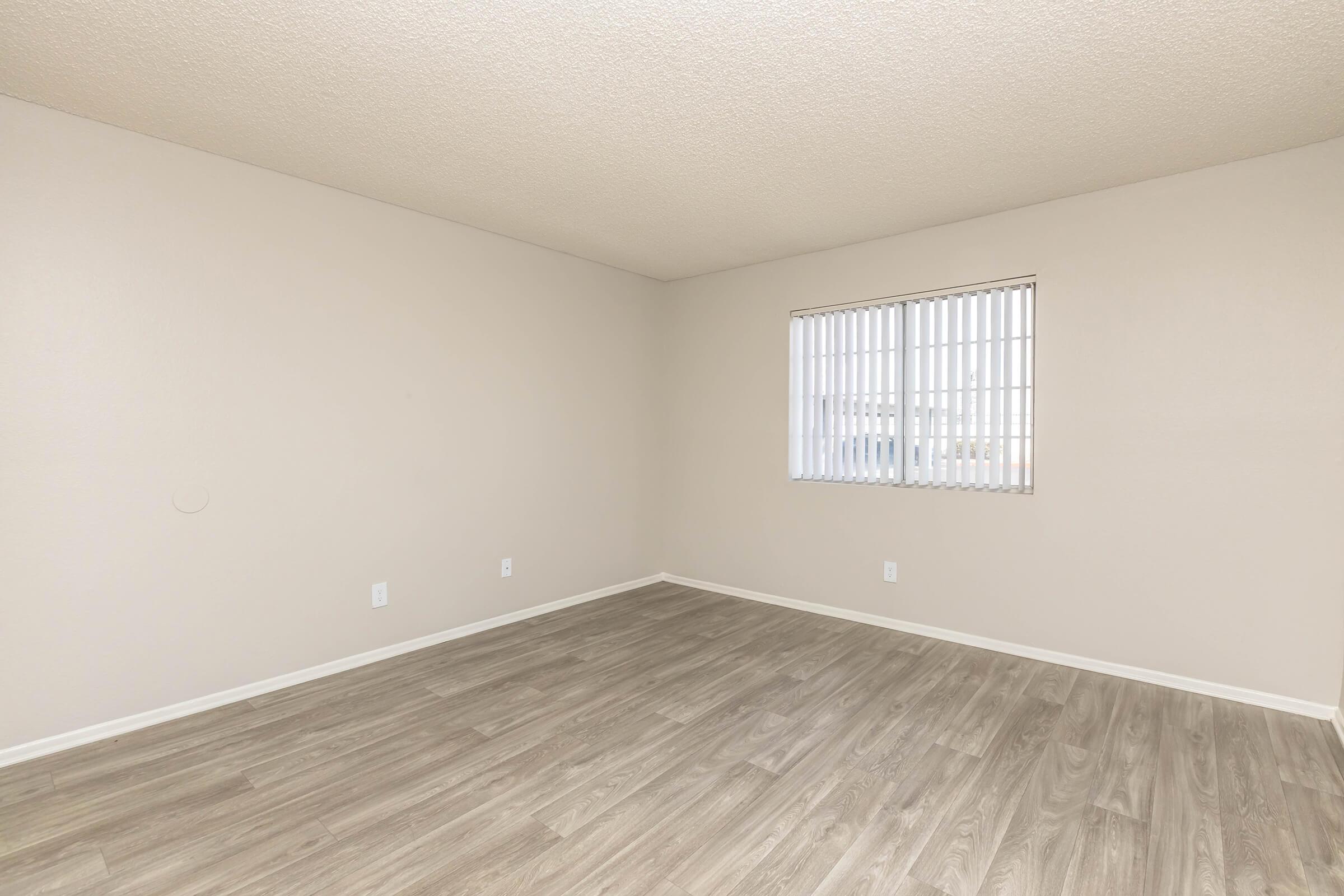 Empty room with light-colored walls and a textured ceiling. The floor is covered in light wood-like laminate. A window with vertical blinds allows natural light to enter. No furniture or decorations are present, giving the space a clean and spacious appearance.