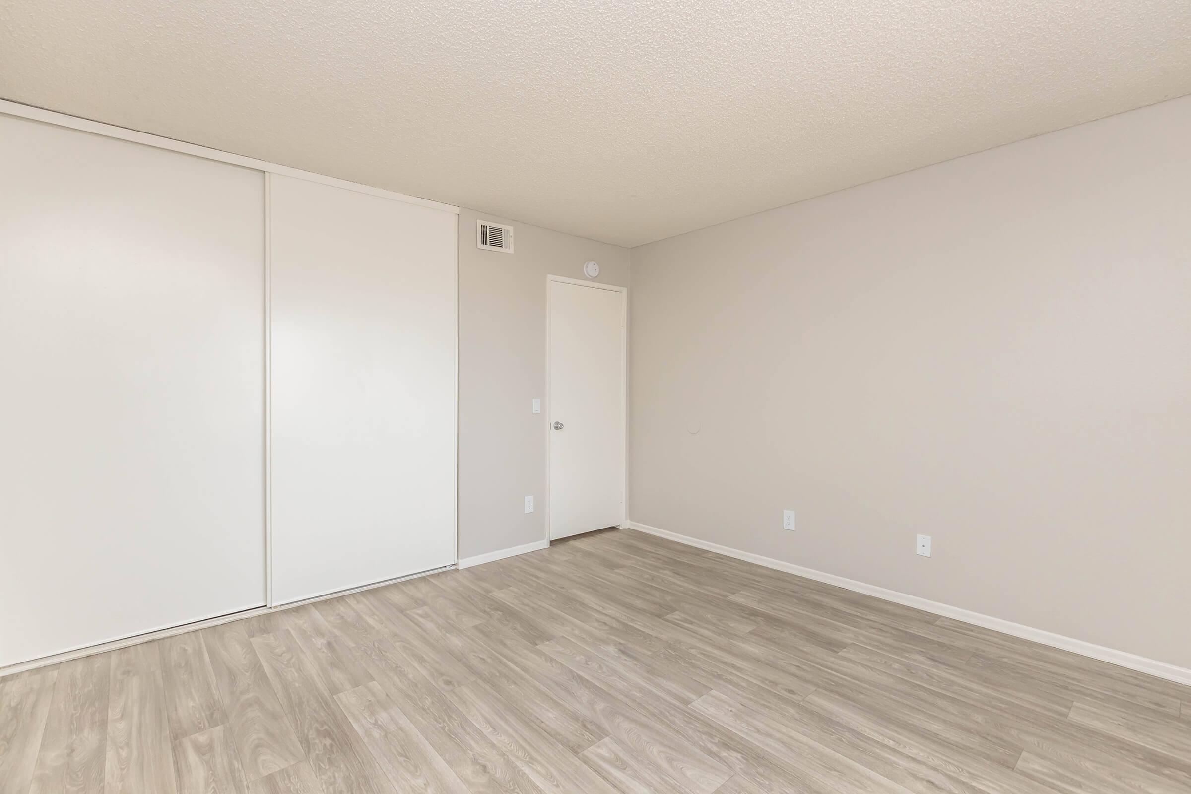 A spacious, empty room with light-colored walls and a wood-like floor. The room features a sliding closet door on the left and a standard door on the right, both of which are closed. Natural light fills the space, creating a bright and airy atmosphere.