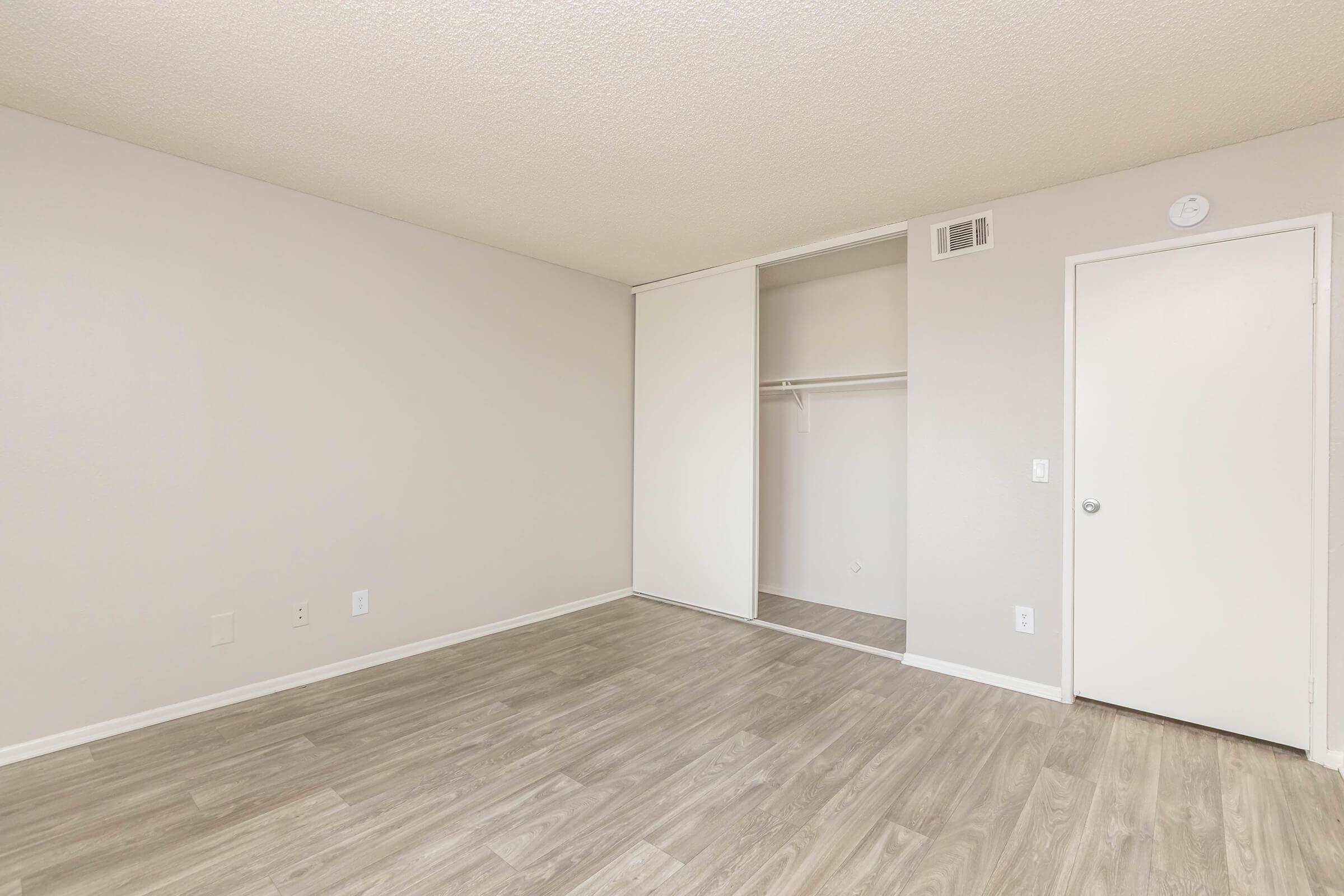 A spacious, empty room with light-colored walls and a textured ceiling. The floor is covered with light-colored laminate or hardwood. On one side, there’s a closet with sliding doors, and a plain white door leads to another area. Natural light illuminates the space, creating a bright and open feel.