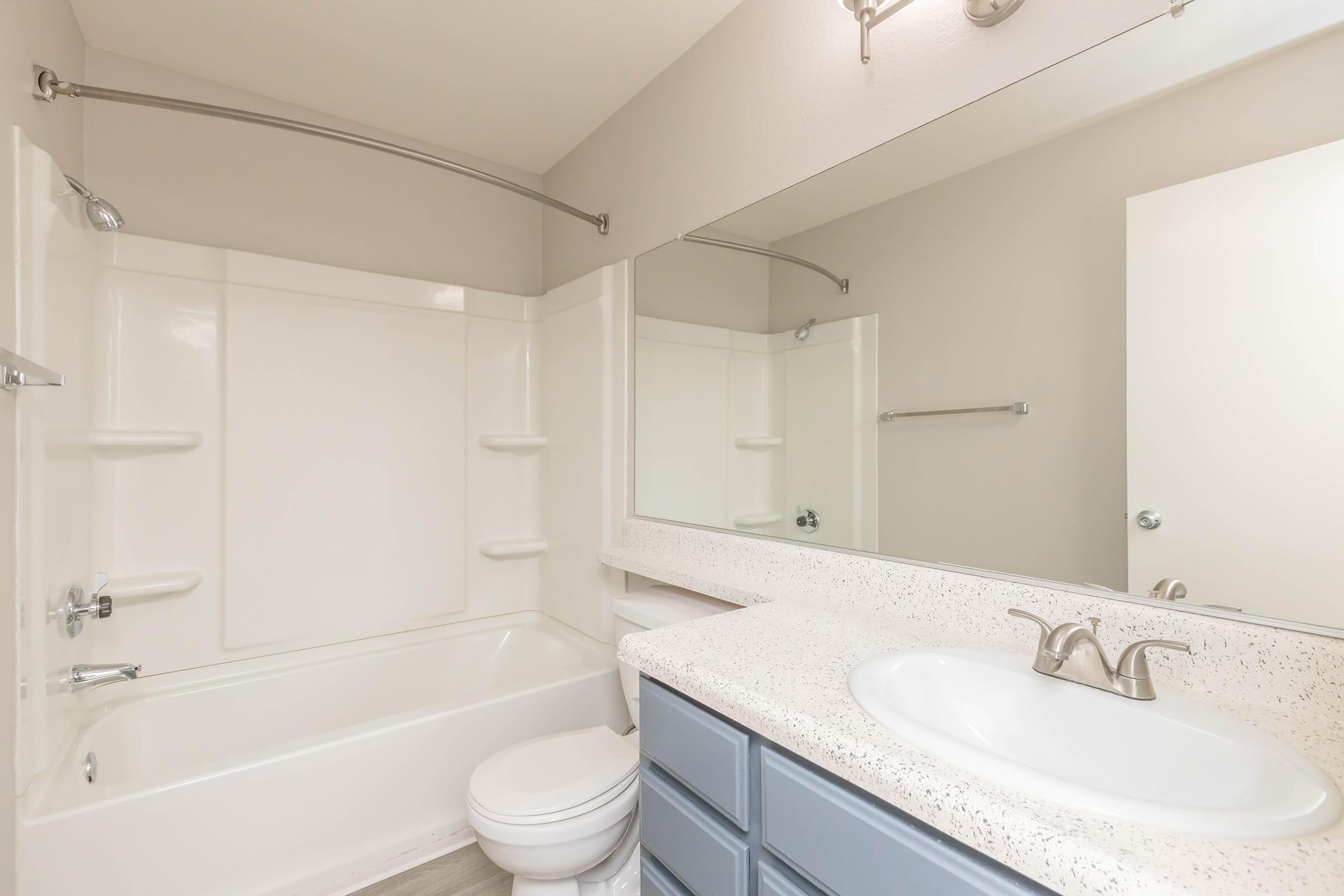 A clean modern bathroom featuring a bathtub with a shower, a white sink with a stylish faucet, and a long countertop with blue cabinetry. The walls are light beige, and there is a large mirror above the sink. Bright lighting enhances the spacious feel of the room.