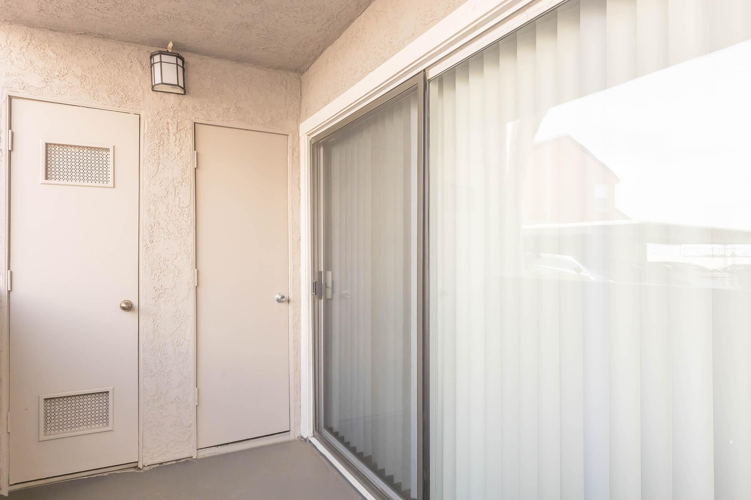 A neutral-colored exterior view of a small balcony or porch area, featuring a sliding glass door with vertical blinds and a plain door on the left, along with a light fixture above. The sunlight creates a bright atmosphere, enhancing the simple yet modern design of the space.