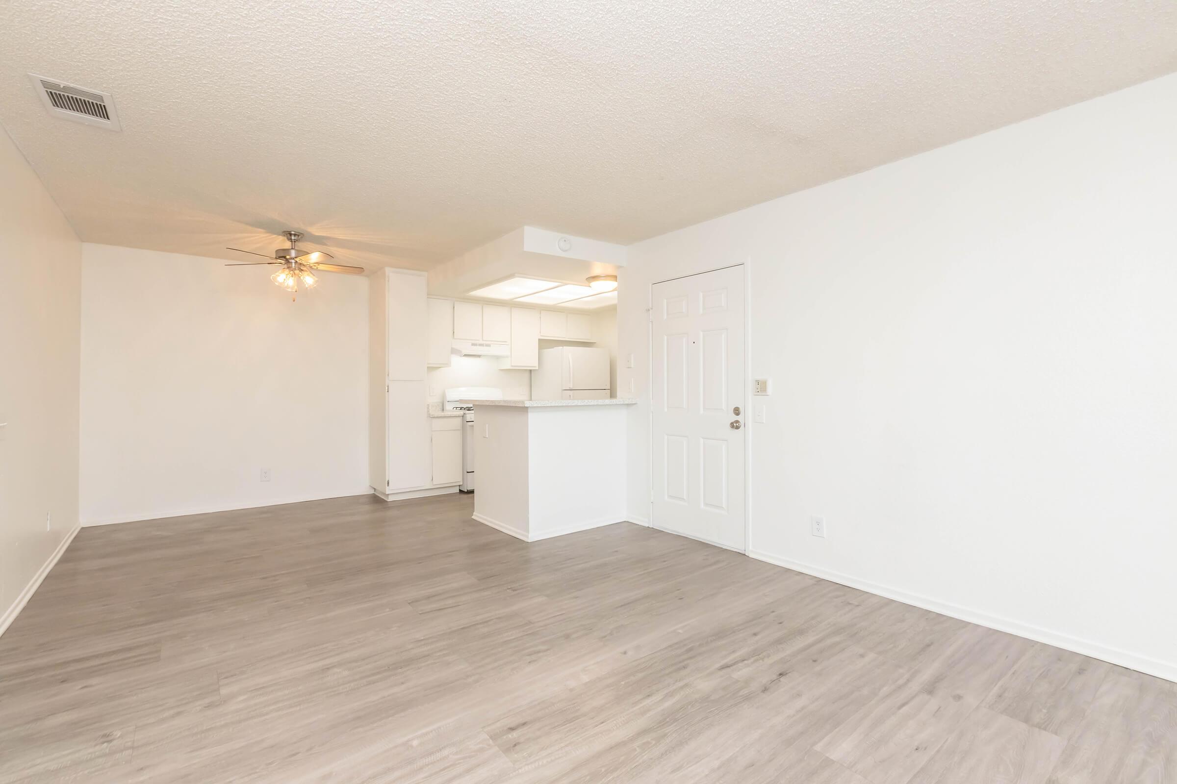 Bright and spacious living area with light-colored walls and wood-like flooring. Includes a ceiling fan, an open kitchen with white cabinets, and a door leading outside. The space appears inviting and well-lit, ideal for comfortable living.