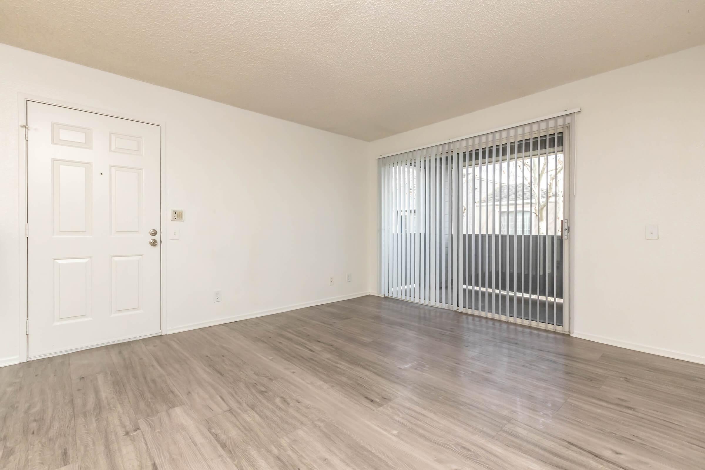 Empty living room with light-colored walls and sleek laminate flooring. A white door is visible on the left, and large sliding glass doors at the back lead to a balcony, partially covered with vertical blinds. The room has a bright and spacious feel due to the natural light coming through the doors.