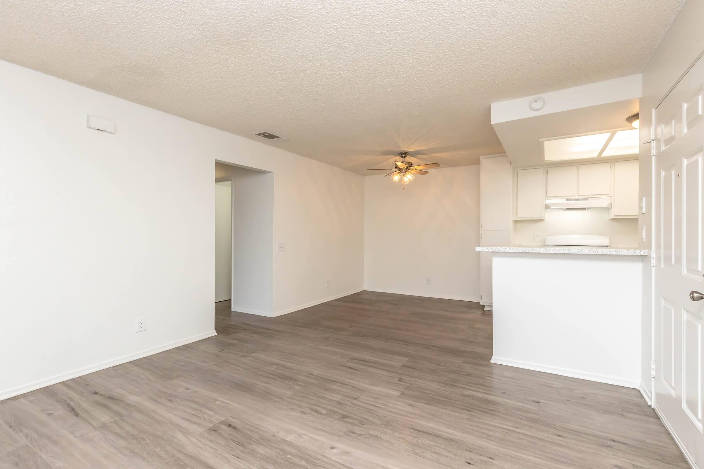Spacious living area featuring light-colored walls and laminate flooring, with a ceiling fan for airflow. An open kitchen with a breakfast bar is visible, along with a doorway leading to another room. Natural light enters through the windows, creating a bright and inviting atmosphere.