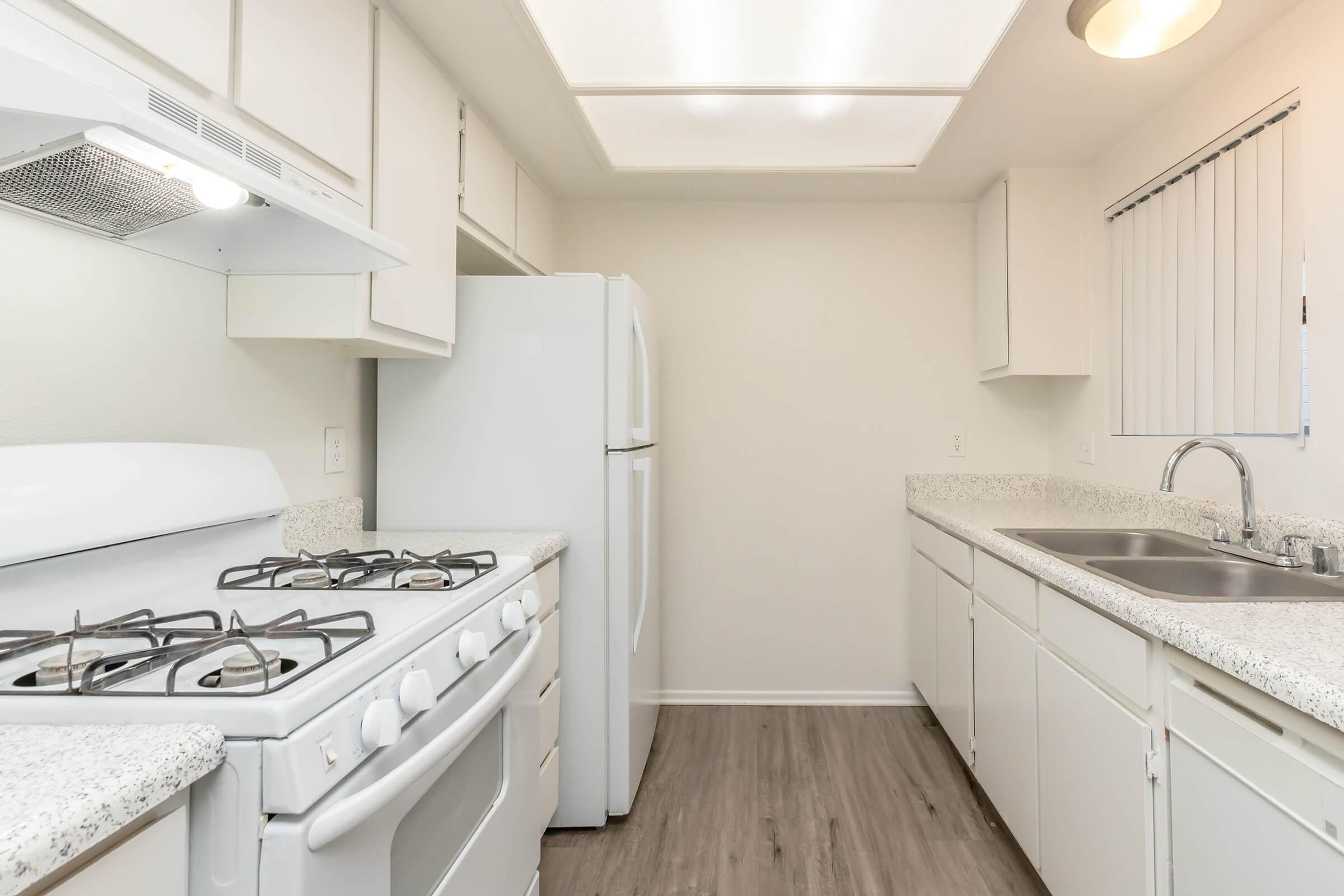 A modern kitchen featuring white cabinets, a gas stove, and an oven. There is a white refrigerator to the right, and a double sink with a stylish faucet. The countertop is a light speckled design, and the floor has a wooden appearance. Natural light enters through a window with vertical blinds, illuminating the space.