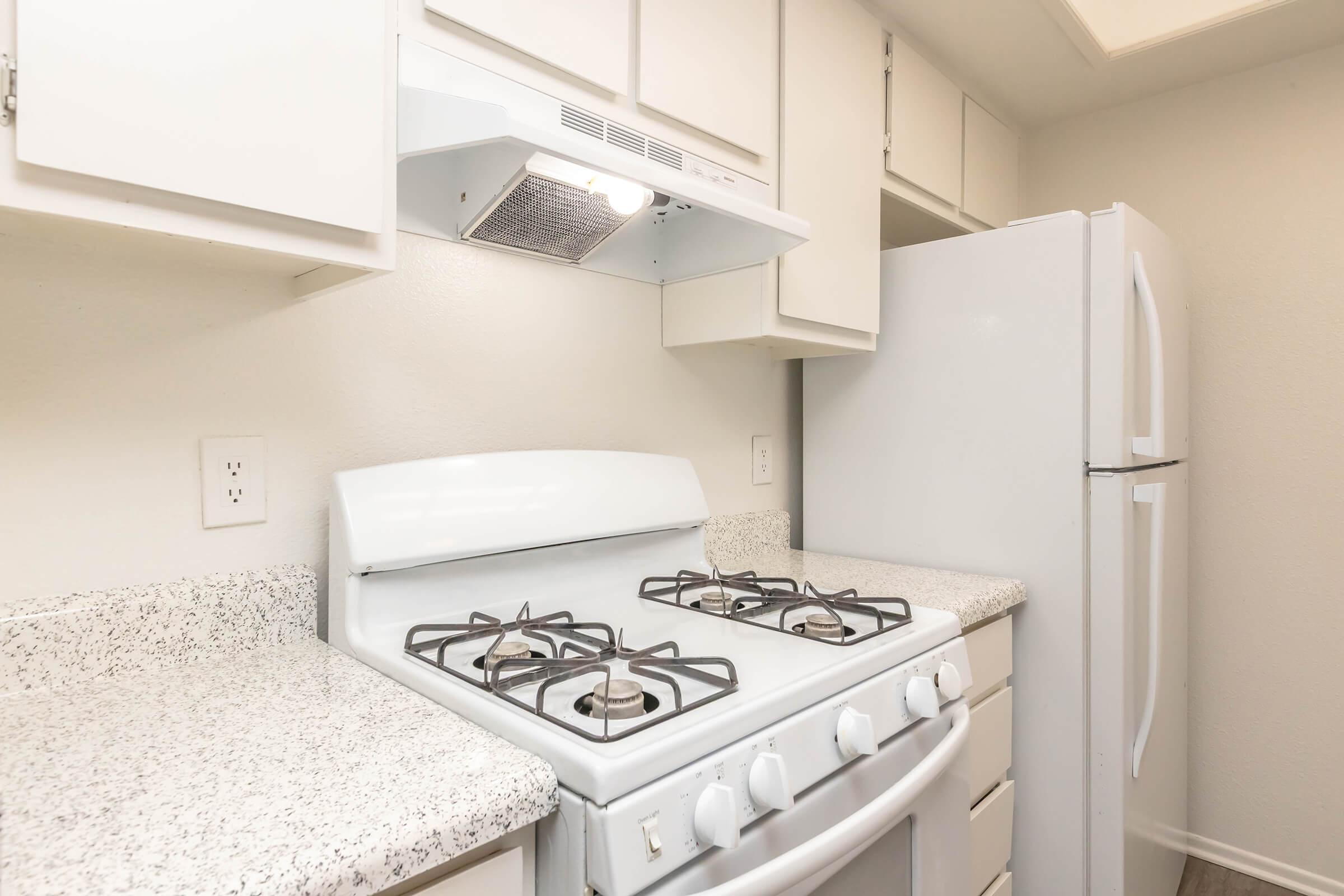 A clean kitchen featuring a white gas stove with four burners, a range hood above, and a side-by-side refrigerator. The countertops are speckled gray and white, and there are white cabinets above and below. The overall color scheme is light and neutral, creating a bright and airy atmosphere.