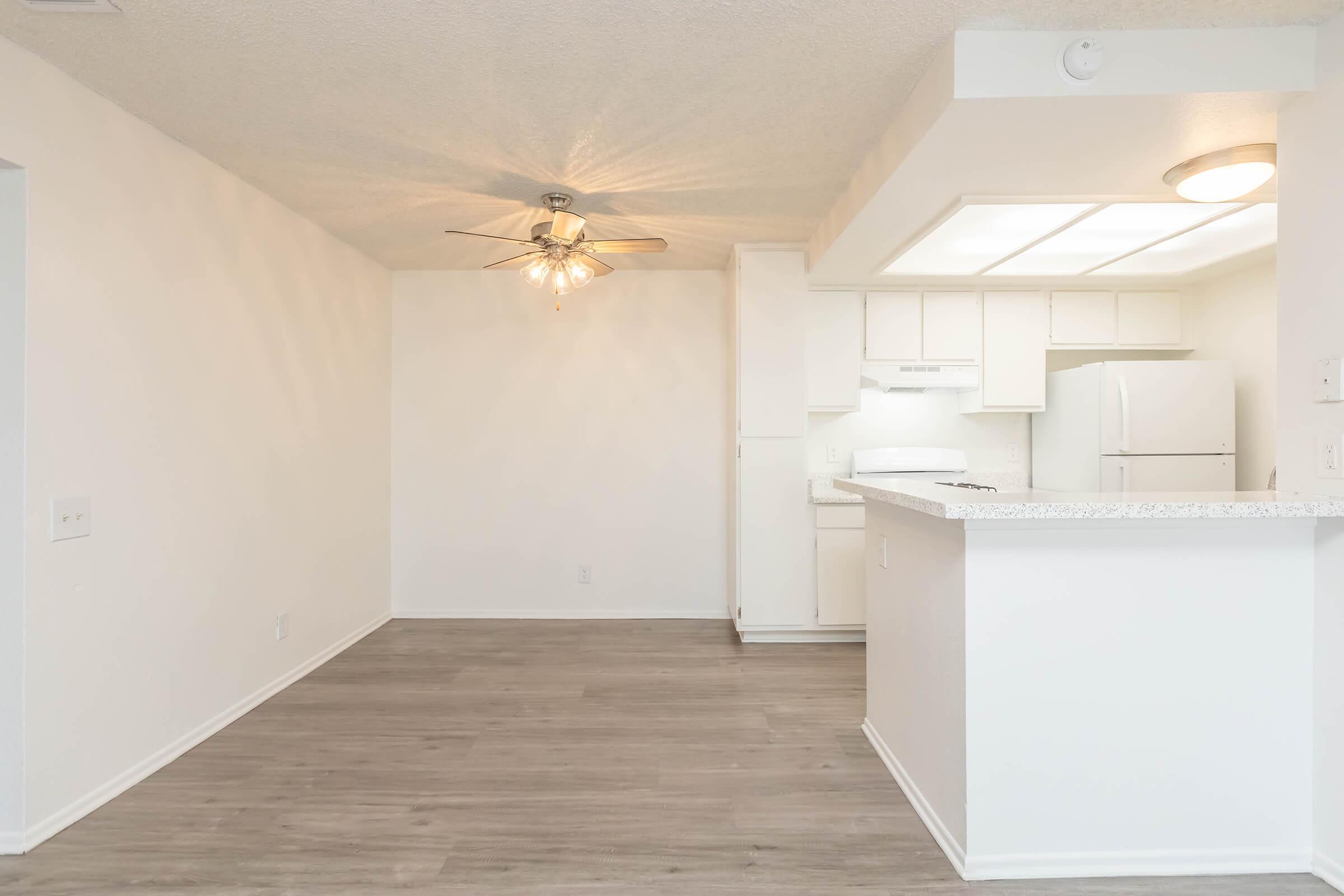 A bright and spacious living area featuring light-colored walls and wood-like flooring. In the background, a kitchen area with white cabinets and appliances is visible, along with a ceiling fan that adds a cozy touch. The layout is open, creating a welcoming atmosphere.