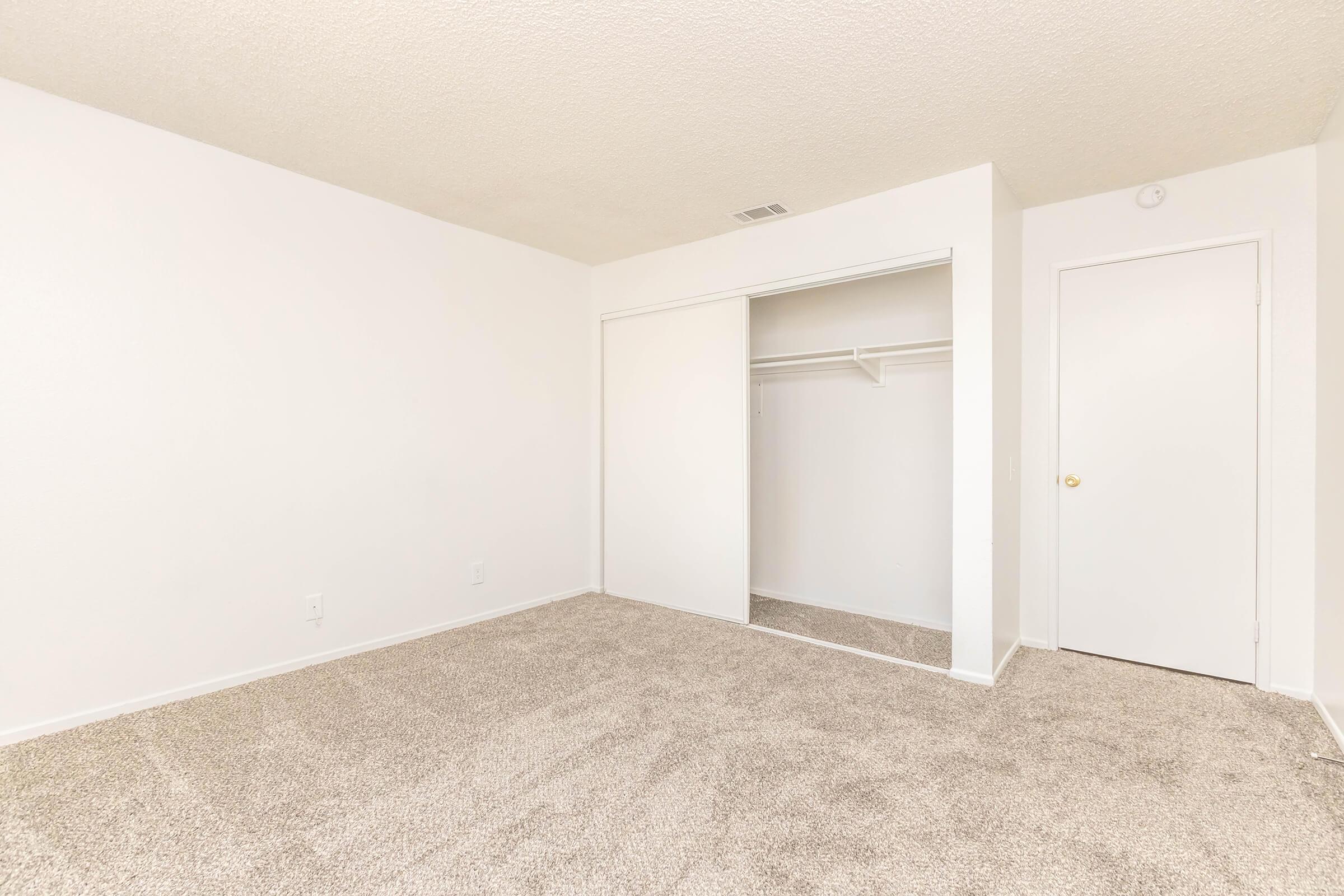 A spacious, empty room with light beige carpet and white walls. A closet with sliding doors is visible on the left side, and a plain door is seen on the right. The room is well-lit, creating an airy feel with no furniture present.