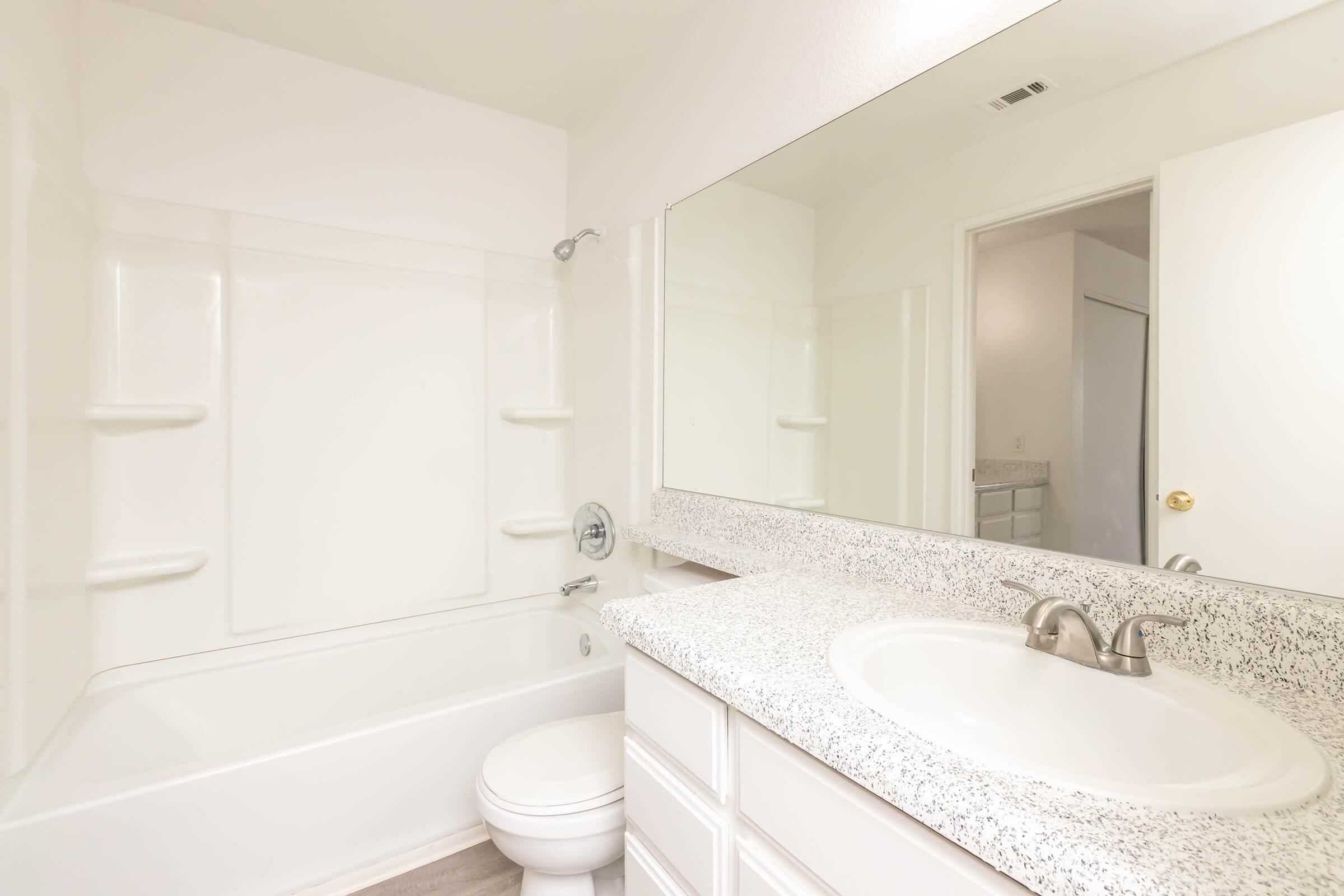 A clean, modern bathroom featuring a white bathtub and shower combination, a large mirror above a granite countertop with a sink, and a toilet. The walls are painted in light colors, enhancing the bright and airy feel of the space. A partially open door leads to an adjoining area.