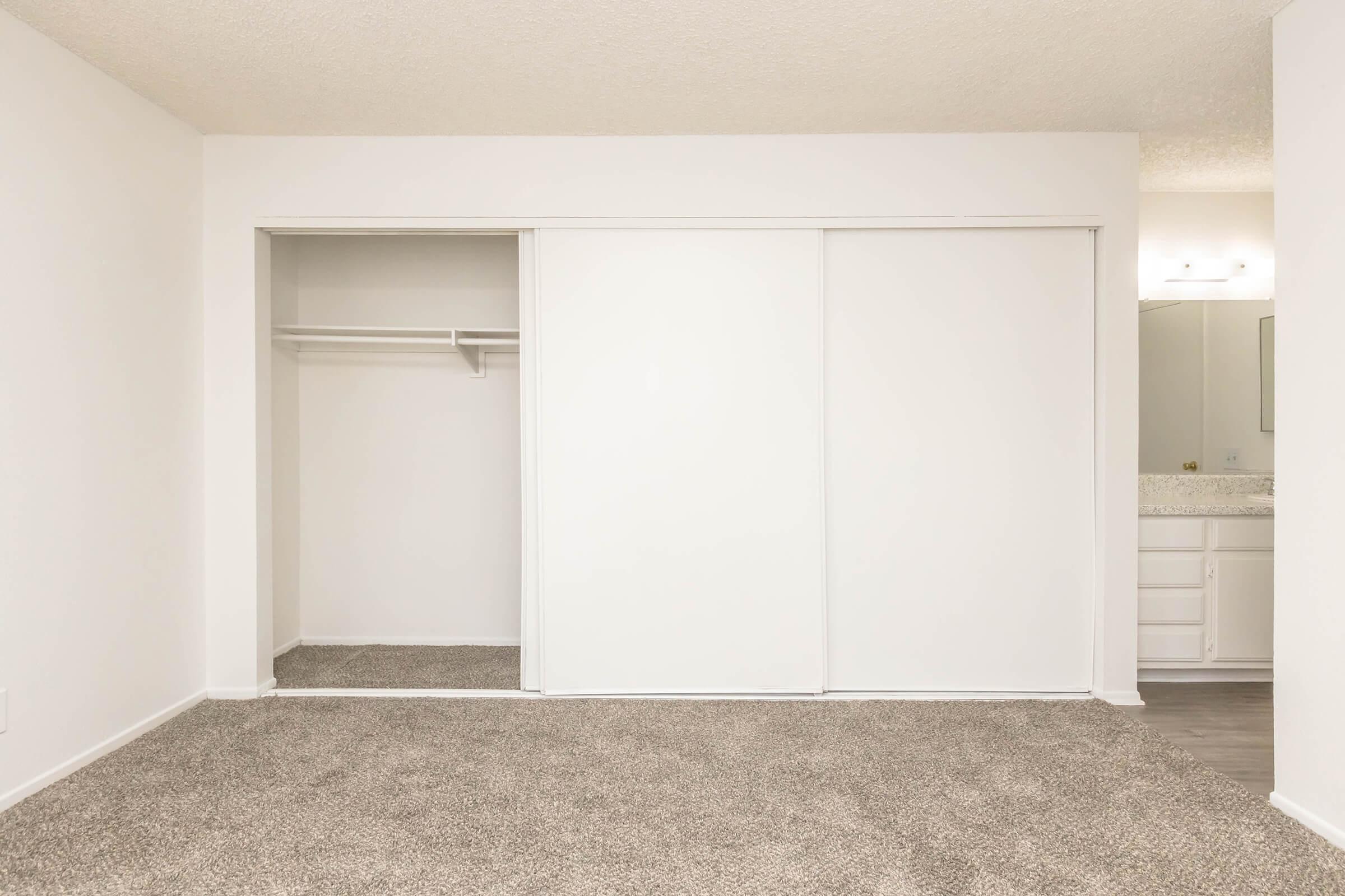 A spacious, empty room featuring a closet with sliding white doors. The floor is covered in light beige carpet, and there is a bathroom area visible in the background with a mirror and countertop. The walls are painted white, creating a bright and airy atmosphere.