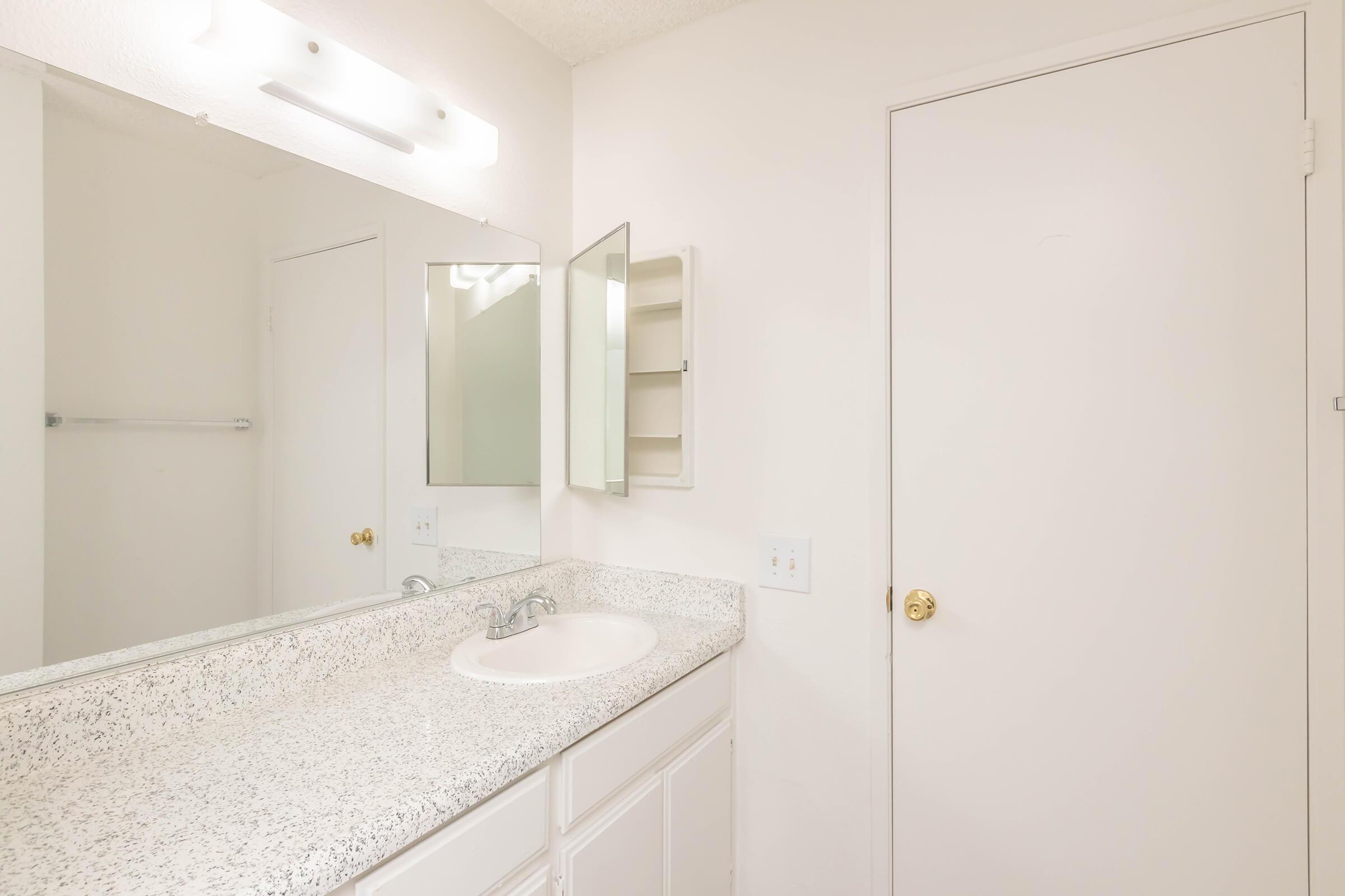 A well-lit, clean bathroom featuring a white countertop with a sink, a mirrored cabinet, and a door leading to another room. The walls are painted white, and there is a light fixture above the mirror. The overall decor is simple and modern.