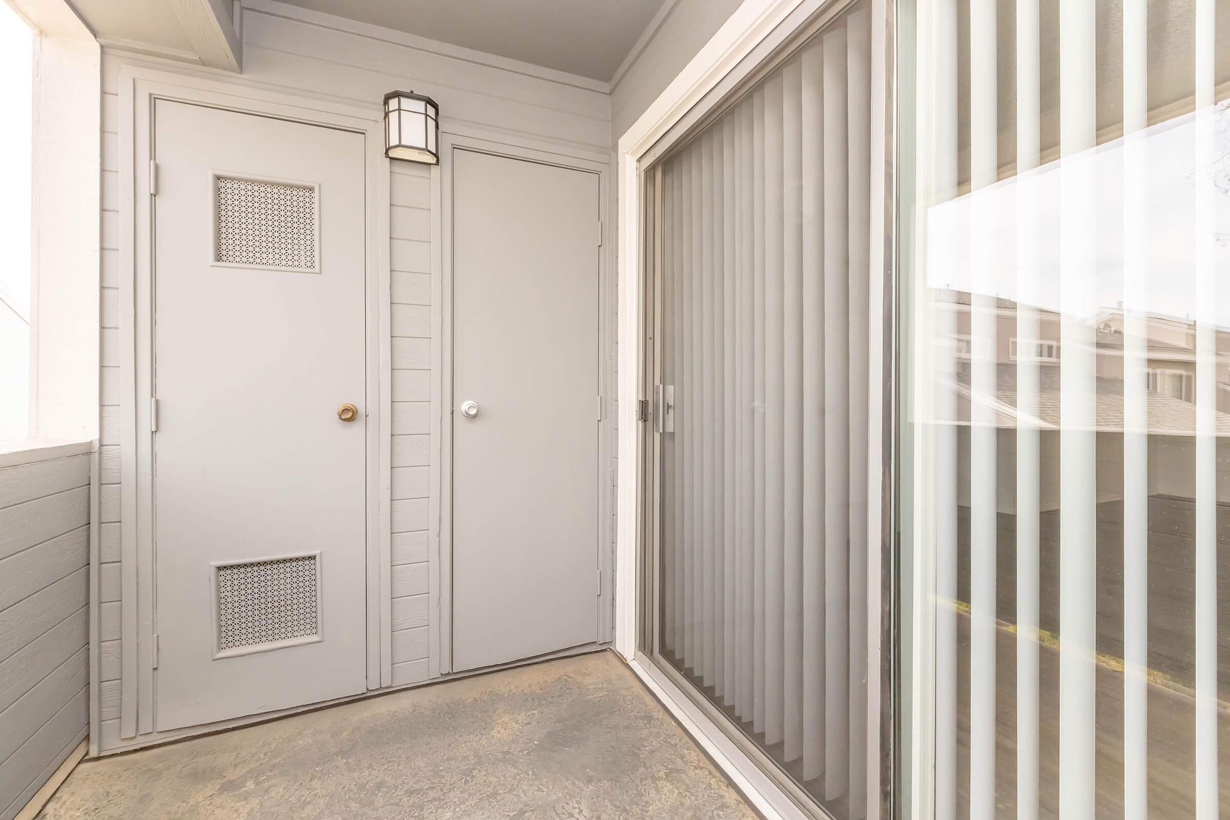 A small balcony area featuring a gray door with a mesh panel, a round light fixture on the wall, and a sliding glass door with vertical blinds. The floor is concrete, and the walls are light gray with horizontal wooden panels. The view is partially obstructed by the sliding door.