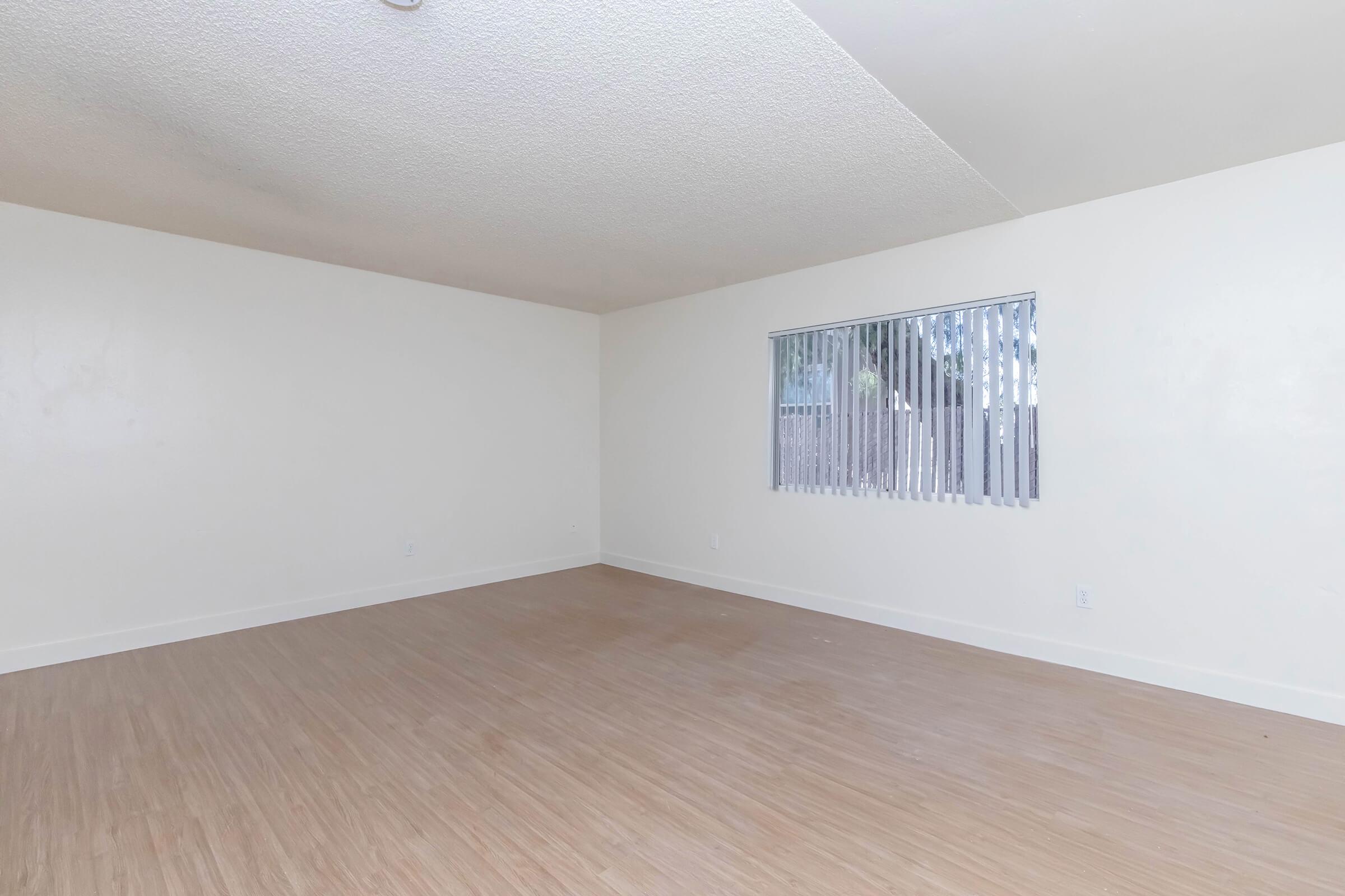 Spacious empty room with light-colored walls and a textured ceiling. A window with vertical blinds allows natural light to enter. The floor is light wood, and there are no furnishings or decorations, creating a clean and minimalistic atmosphere.