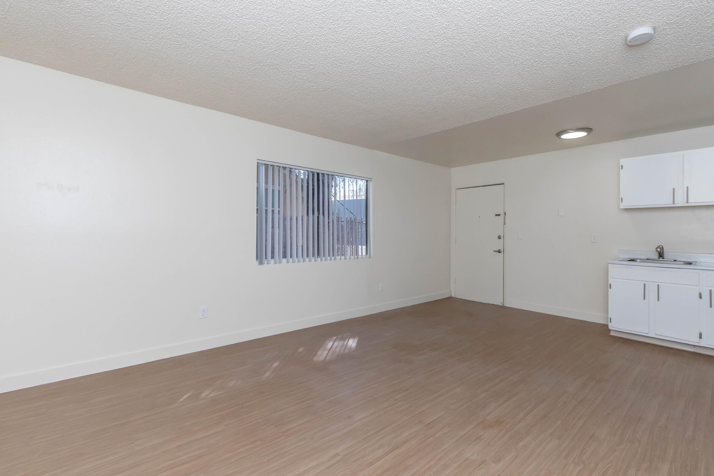 A spacious, empty room with beige walls and light wood flooring. There's a window with vertical blinds on the left, allowing natural light in. The entrance door is on the right side, and a small kitchenette is visible against the back wall, featuring white cabinets and a sink. The overall atmosphere is bright and clean.