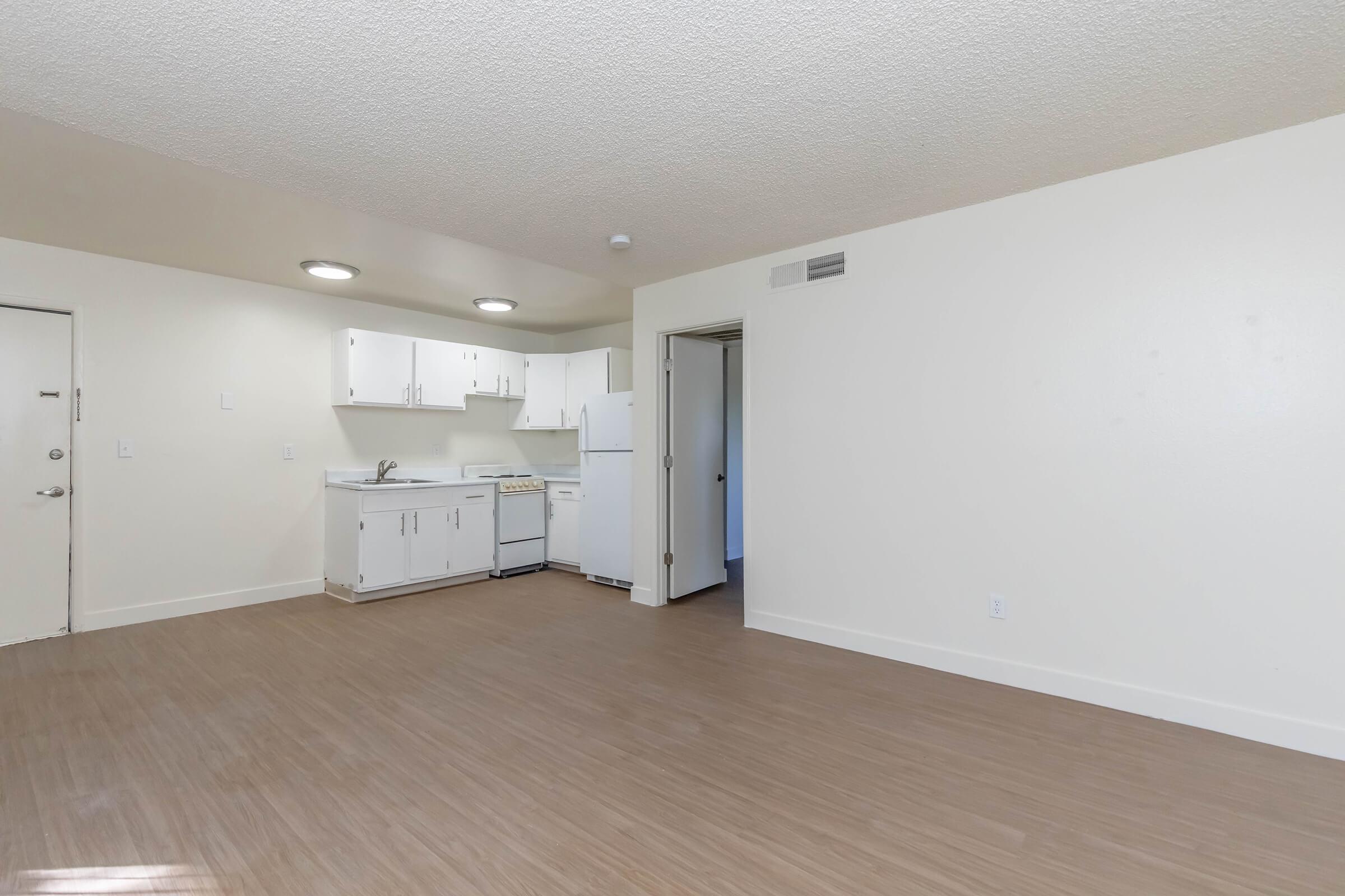 A spacious, modern apartment interior featuring light-colored walls and laminate flooring. The kitchen area has white cabinets, a stove, and a refrigerator. An open layout connects the kitchen to the living area, with a door leading to another room and a front door visible. Bright, natural light fills the space.