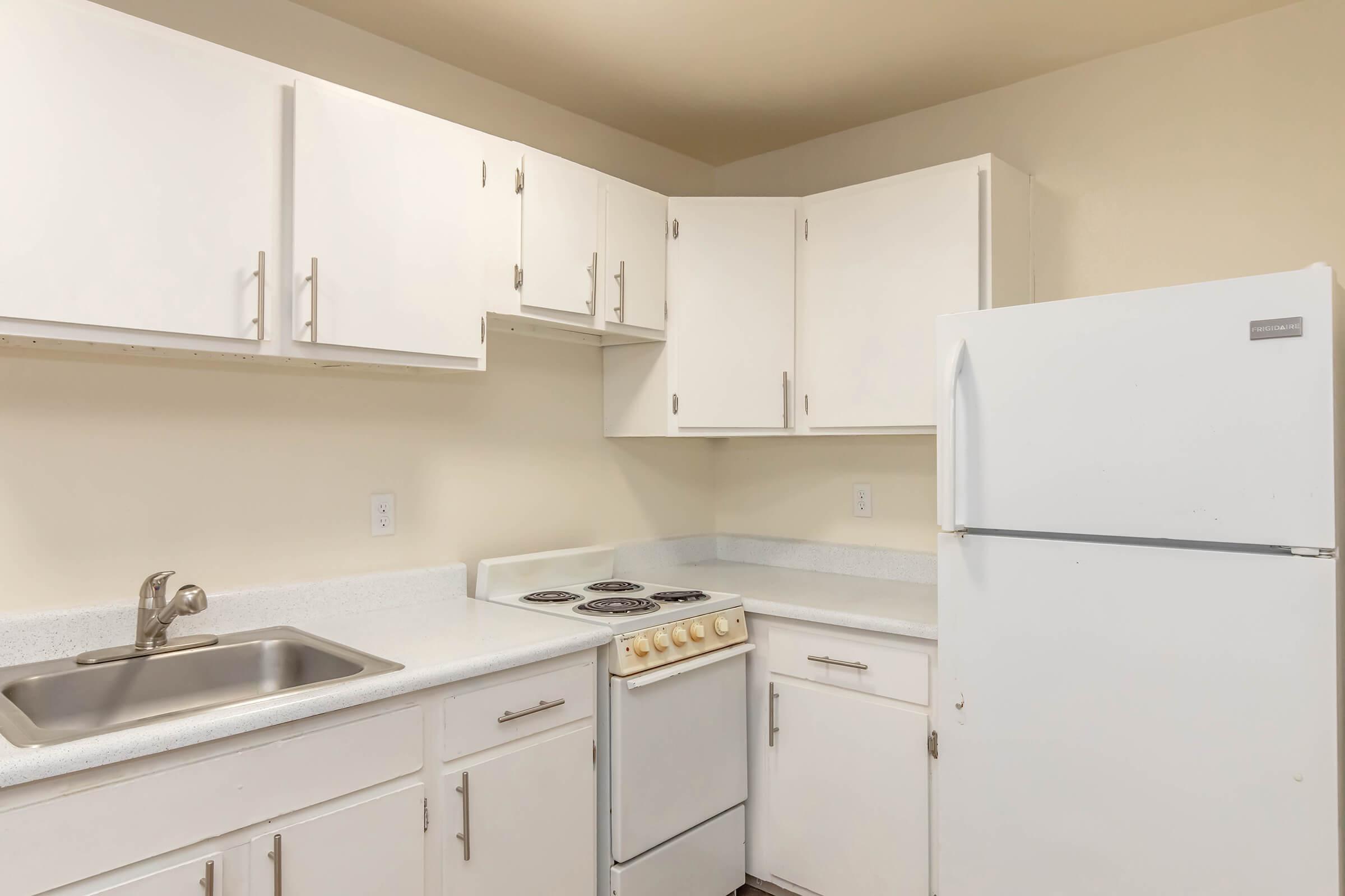 A compact, clean kitchen featuring white cabinets, a sink, a stove with an oven, and a refrigerator. The countertops are light-colored, providing a simple and functional design. The walls are painted in a neutral shade, creating a bright and inviting atmosphere.