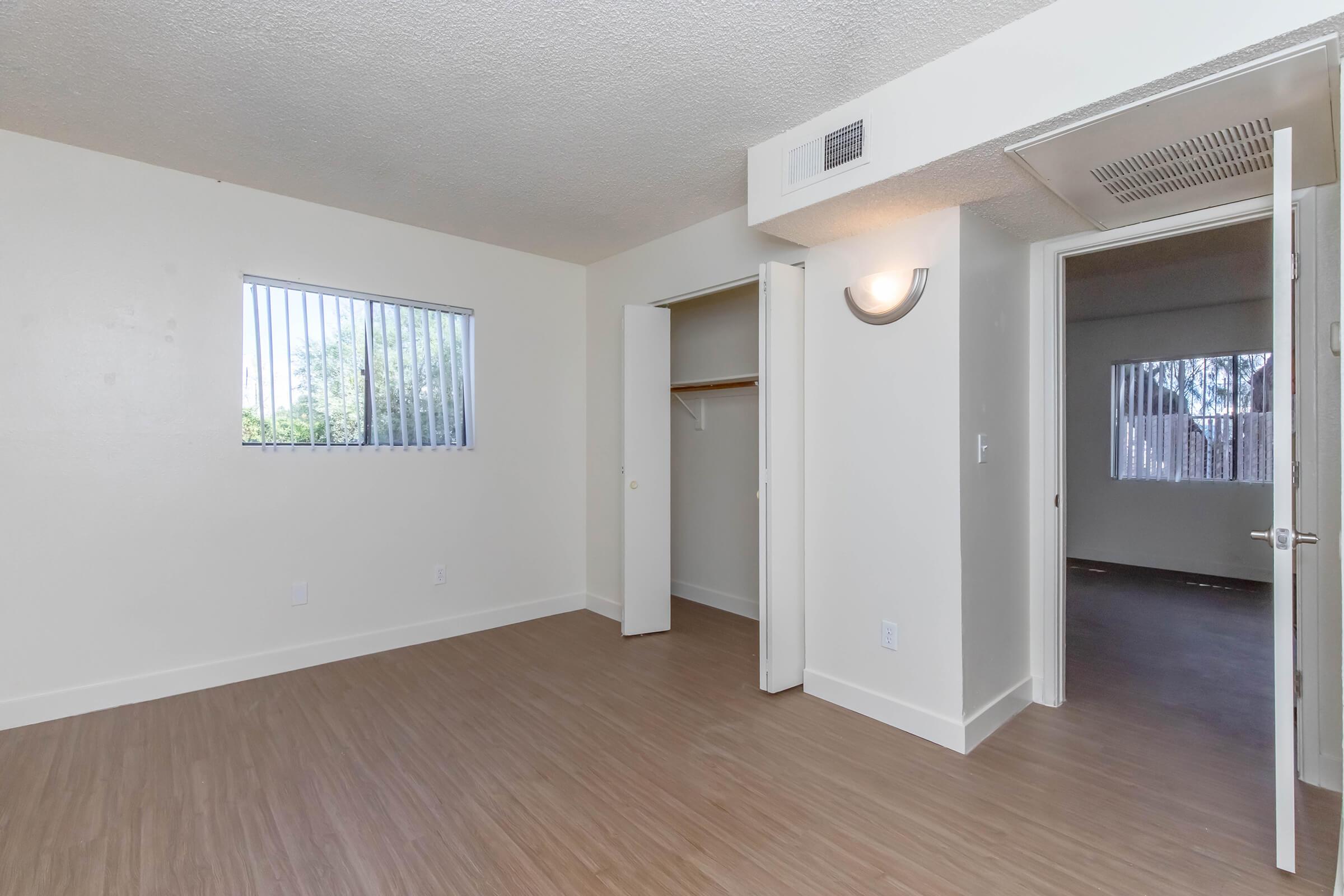 A spacious room with light-colored walls and wood-style flooring. It features a window with blinds, a built-in closet with sliding doors, and a doorway leading to another room. The overall ambiance is bright and minimalistic, suitable for various furniture arrangements.