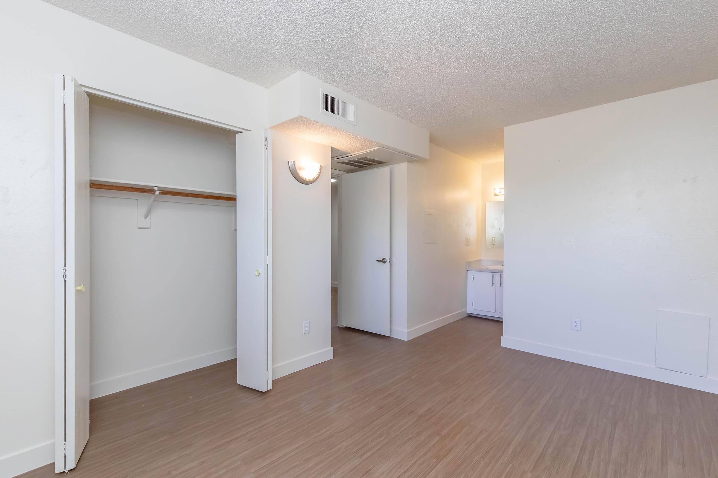 An interior view of a room featuring light-colored walls, laminate flooring, and a closet with an open door. There is a bathroom visible in the background with a vanity and mirror. The space is well-lit and appears modern and inviting.