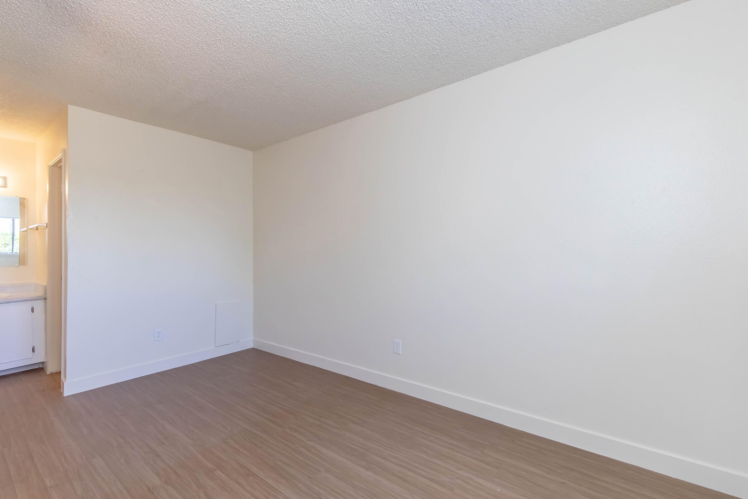 A clean, empty room with light-colored walls and a smooth wooden floor. To the left, there is a doorway leading to a bathroom area, while the right side features a blank wall. Natural light enters from an unseen window, creating a bright ambiance. The space is minimal and ready for decoration or furniture.