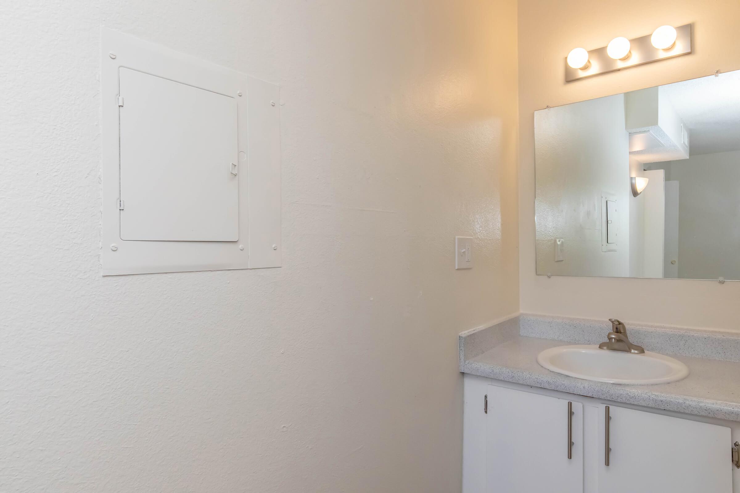A clean bathroom featuring a small sink with a faucet, a lighted mirror above, and a wall-mounted electrical panel. The walls are painted in a light color, creating a bright and open atmosphere.