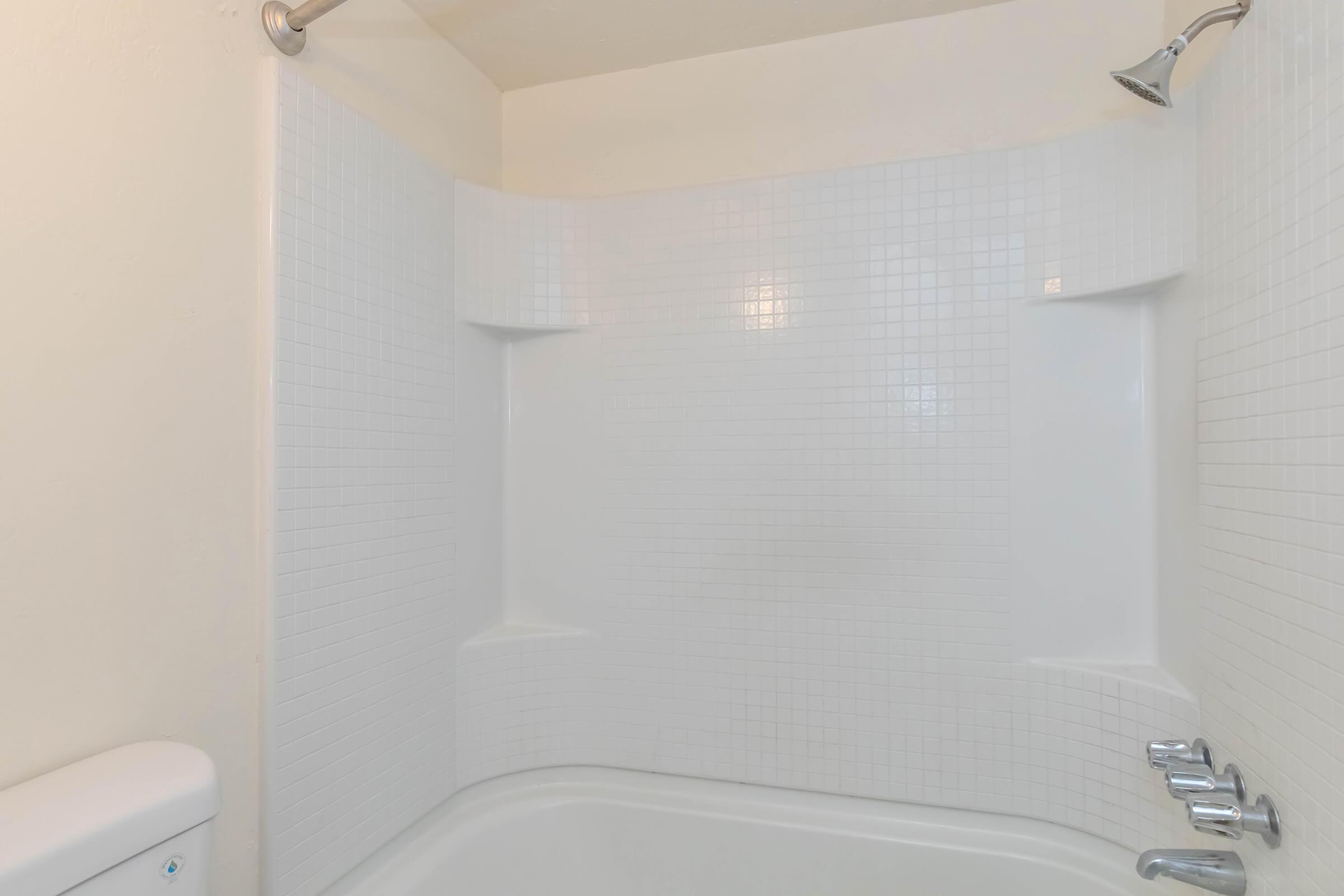 A clean and modern bathroom shower area featuring white tiled walls, a bathtub with a rounded edge, a chrome showerhead, and faucet handles. The space is well-lit with a bright, neutral color scheme.