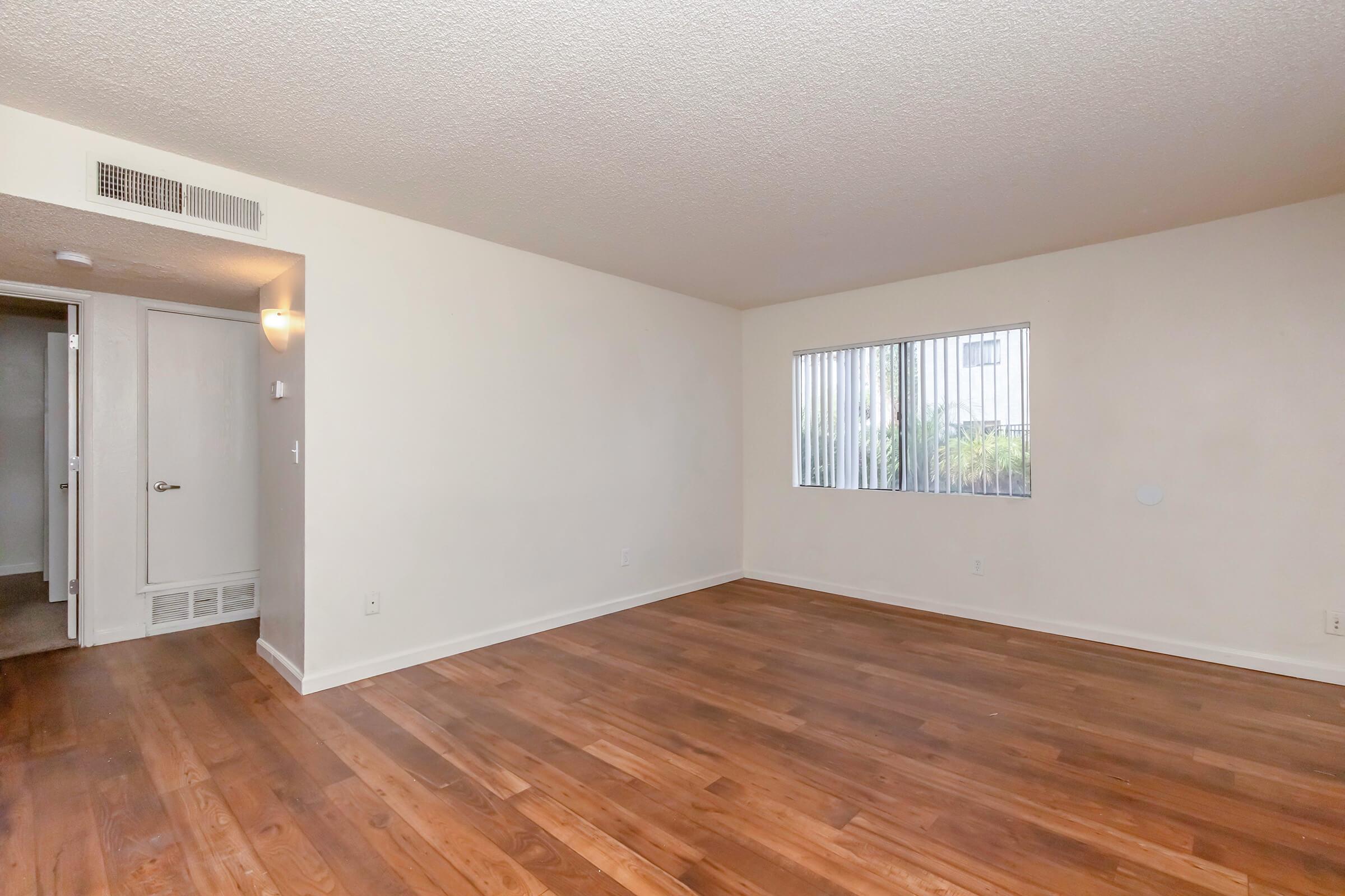 Spacious interior of a vacant room featuring hardwood floors, light-colored walls, and a large window with vertical blinds, providing natural light. A doorway in the background leads to another room, creating an open and inviting atmosphere.