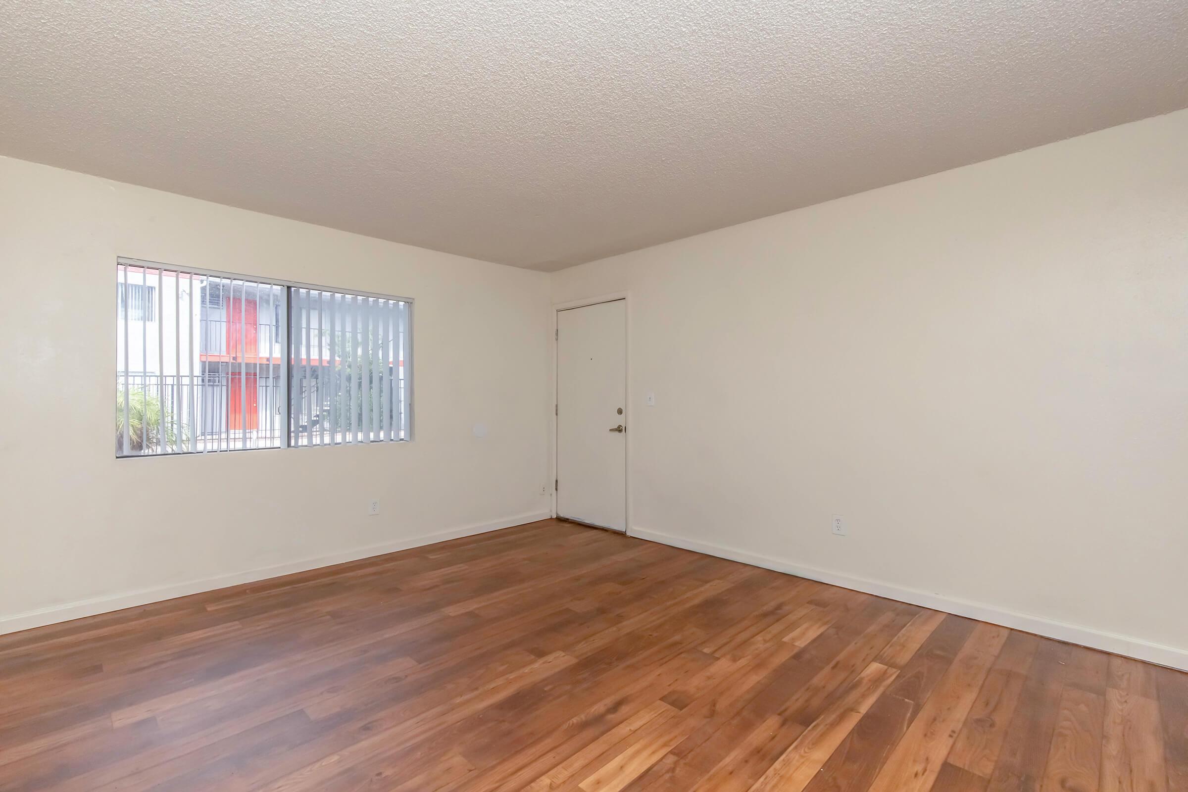 Spacious, empty living room with light-colored walls and a large window. The room features wooden flooring and a door leading outside, offering a bright and airy atmosphere.