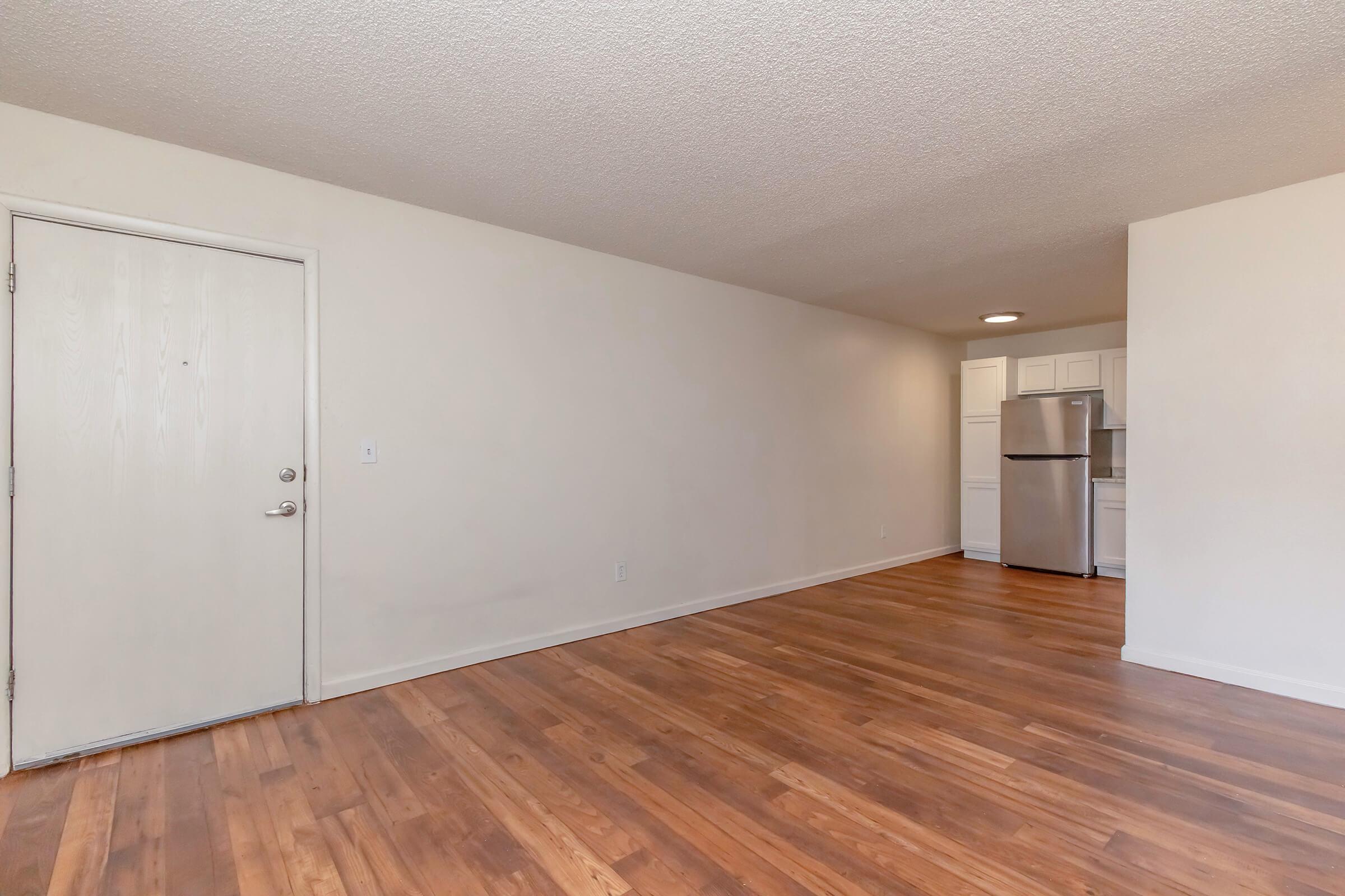 A spacious, empty room with light-colored walls and hardwood flooring. The entrance door is on the left, and a kitchen area with white cabinets and a stainless steel refrigerator is visible in the background. The room is well-lit, creating a bright and inviting atmosphere.