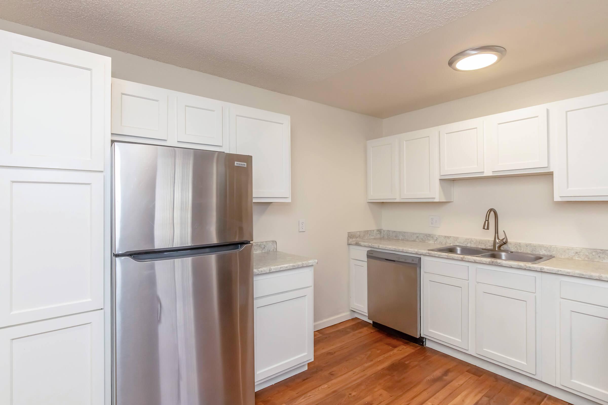 Bright and modern kitchen featuring stainless steel appliances, including a refrigerator and dishwasher. The cabinetry is white with a sleek finish, complemented by a granite countertop. The wood floor adds warmth to the space, while the overhead lighting enhances the inviting atmosphere.