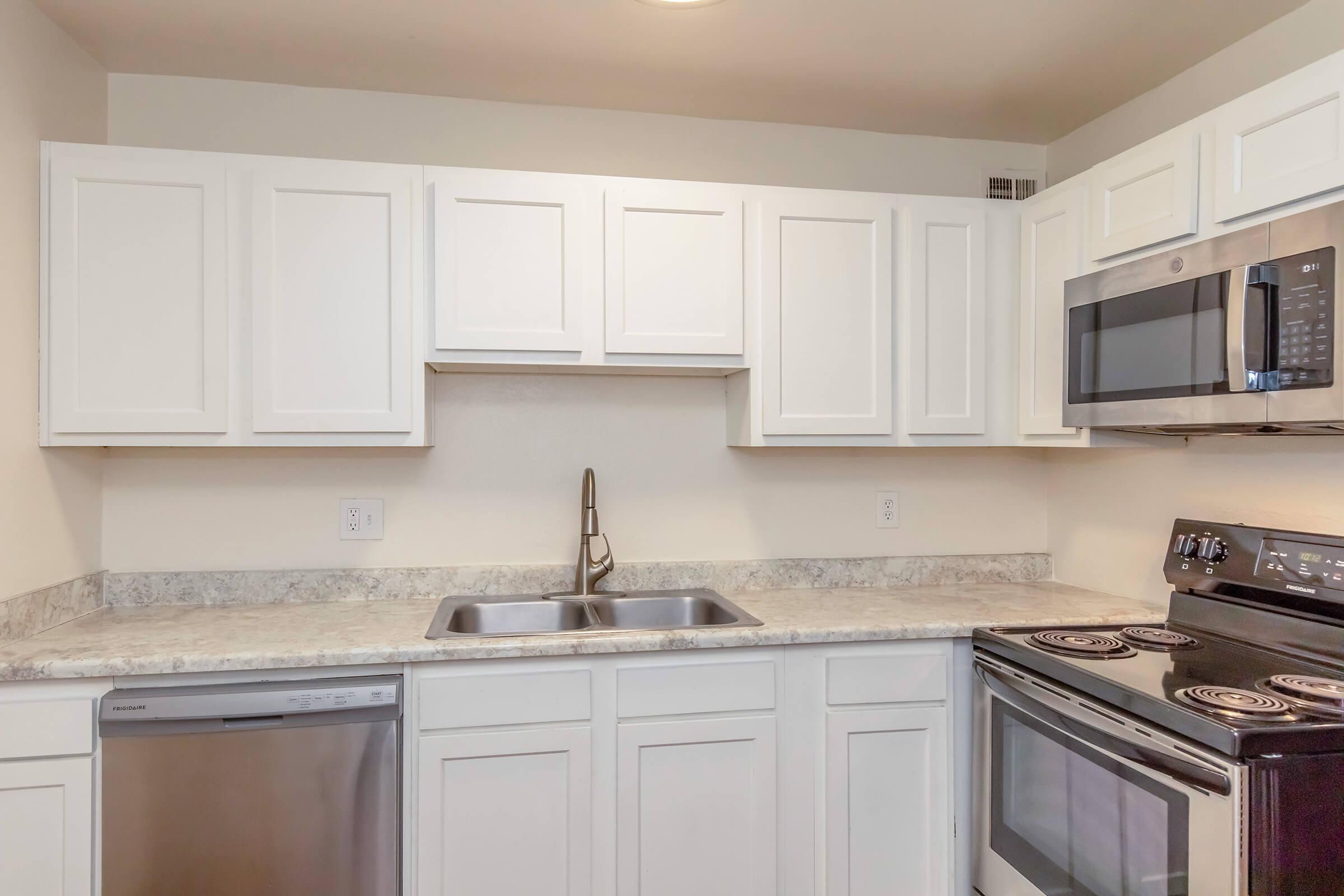 Modern kitchen featuring white cabinetry, a double sink, stainless steel appliances including a microwave, stove, and dishwasher, and a light countertop. The layout is clean and functional, with a neutral color scheme and ample storage space.