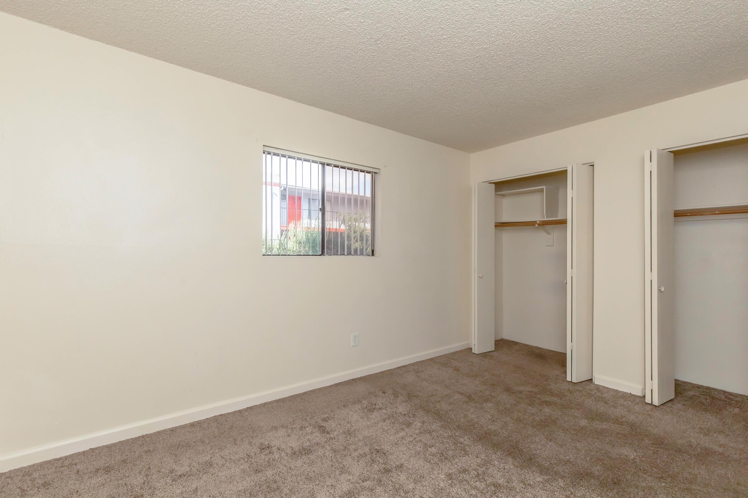 Empty room with beige walls and a textured ceiling. There's a window with vertical blinds on one wall, letting in natural light. Two closet spaces with sliding doors are visible on the opposite wall. The floor is covered in light brown carpet, creating a neutral and open atmosphere.
