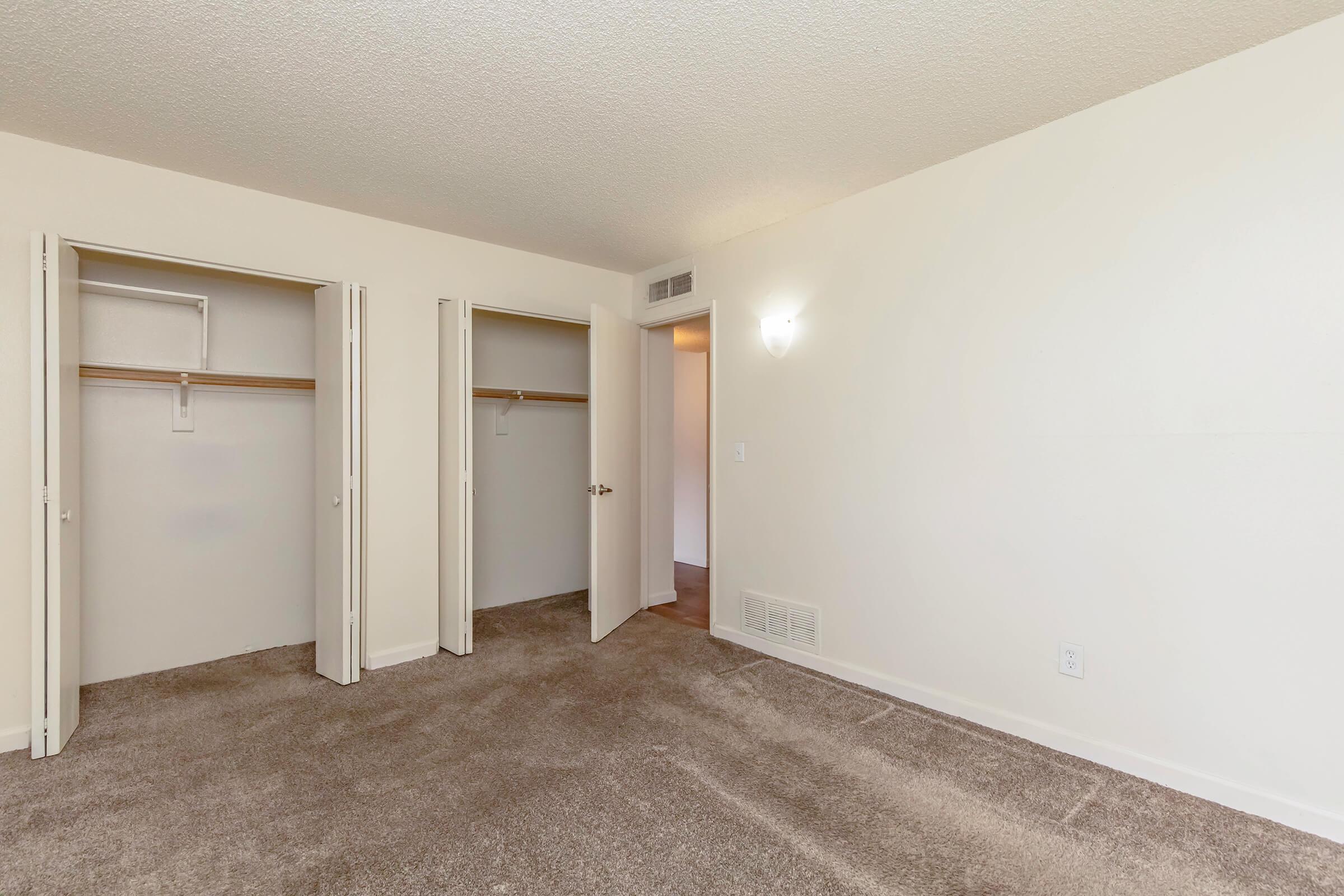 A spacious and empty room featuring light-colored walls and carpeted flooring. There are two open closets on the left side, and a doorway leads to another area of the space. A small wall-mounted light fixture is visible, providing ambient illumination.