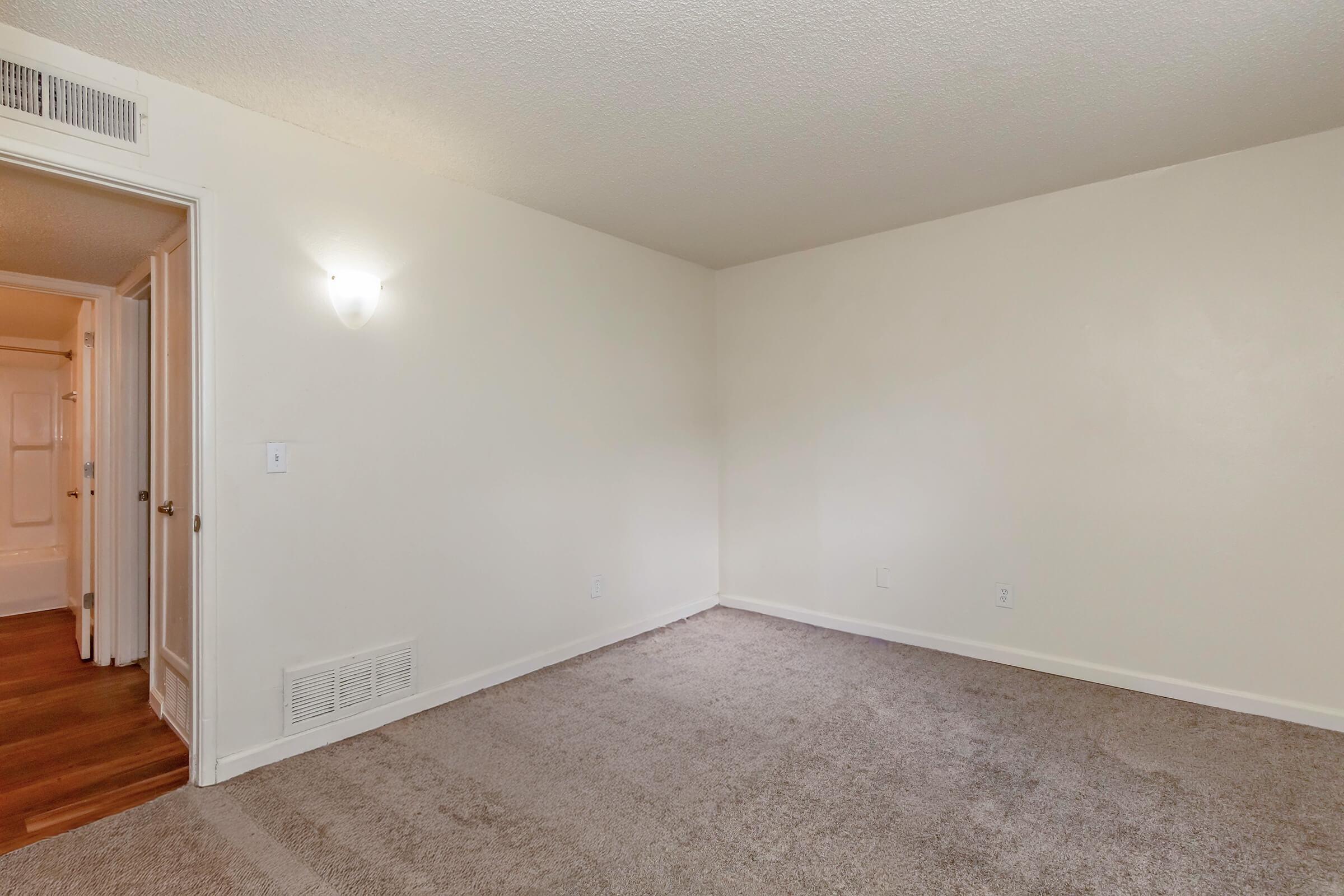 A clean, empty room featuring light-colored walls and beige carpet. There is a wall-mounted light fixture on the left side, and a door leading to a hallway visible in the background. The space appears bright and spacious, ready for furnishing.