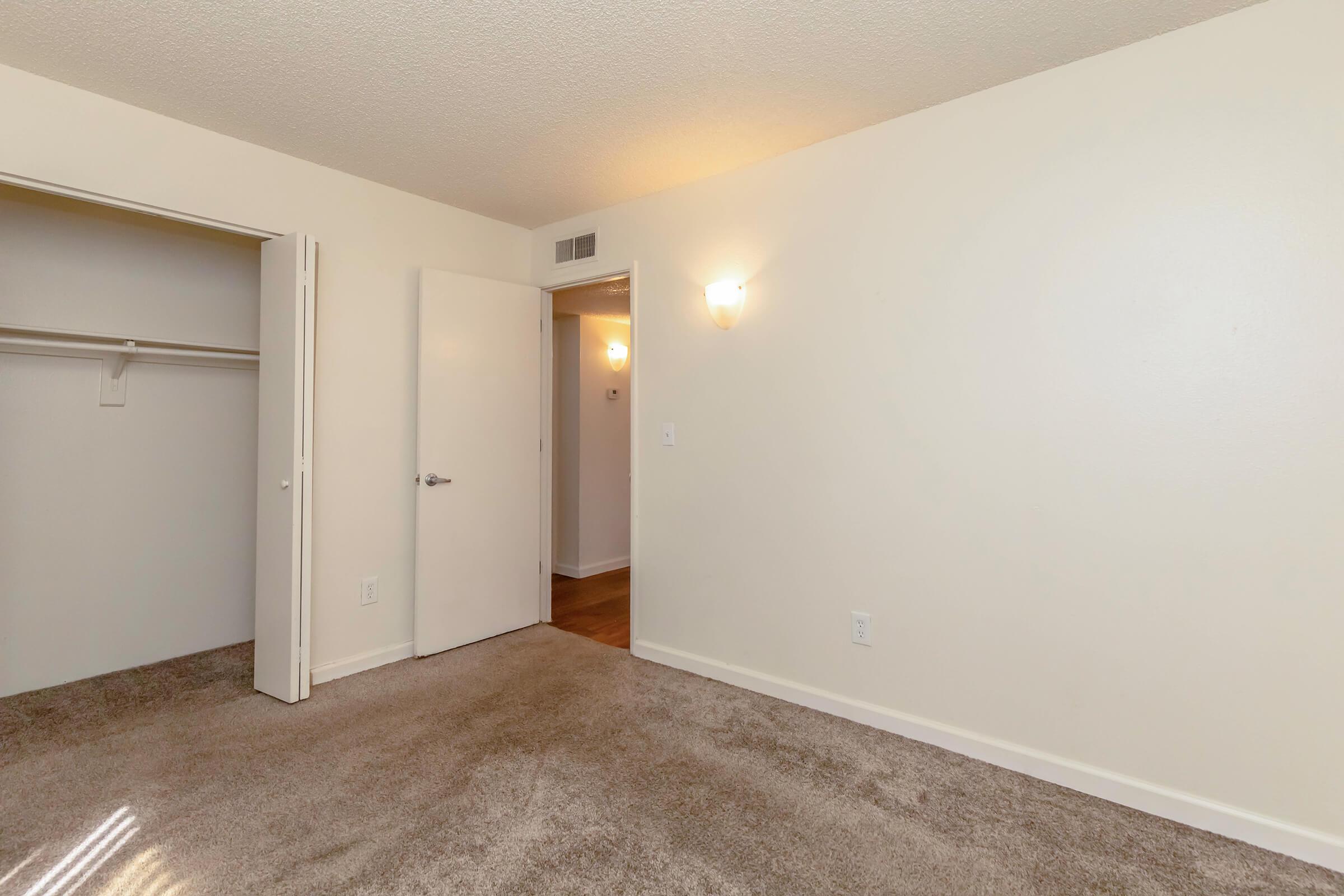 Empty room with light beige walls and carpeted flooring. There is a closet door on the left and a partially open door leading to another space on the right. A light fixture on the wall provides illumination, creating a warm atmosphere in the otherwise unfurnished space.