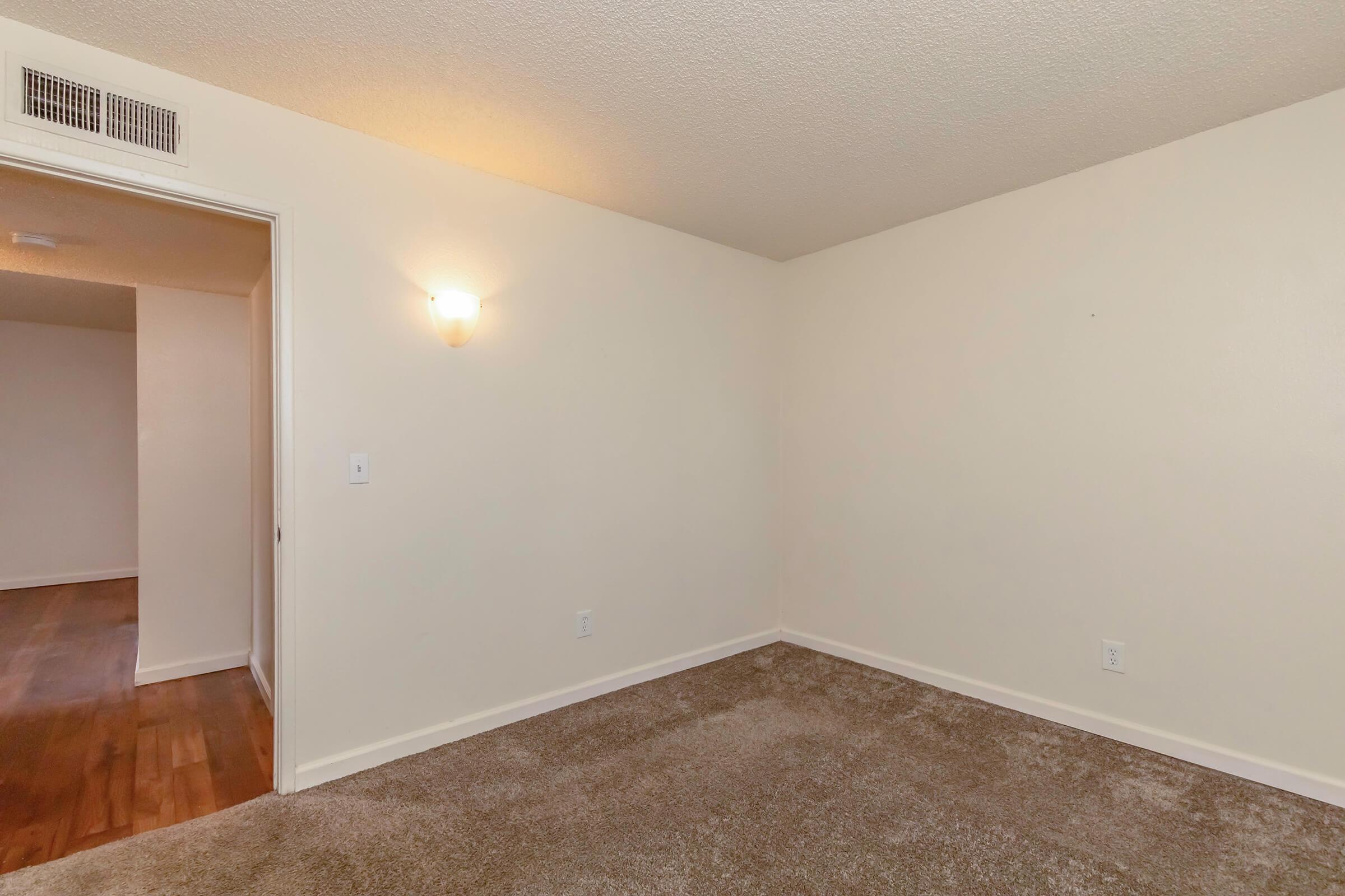 Empty room with light beige walls and a carpeted floor. A light fixture on the wall provides illumination. On one side, there is an open door leading to another room, suggesting a spacious layout. The overall atmosphere is neutral and ready for furnishing.