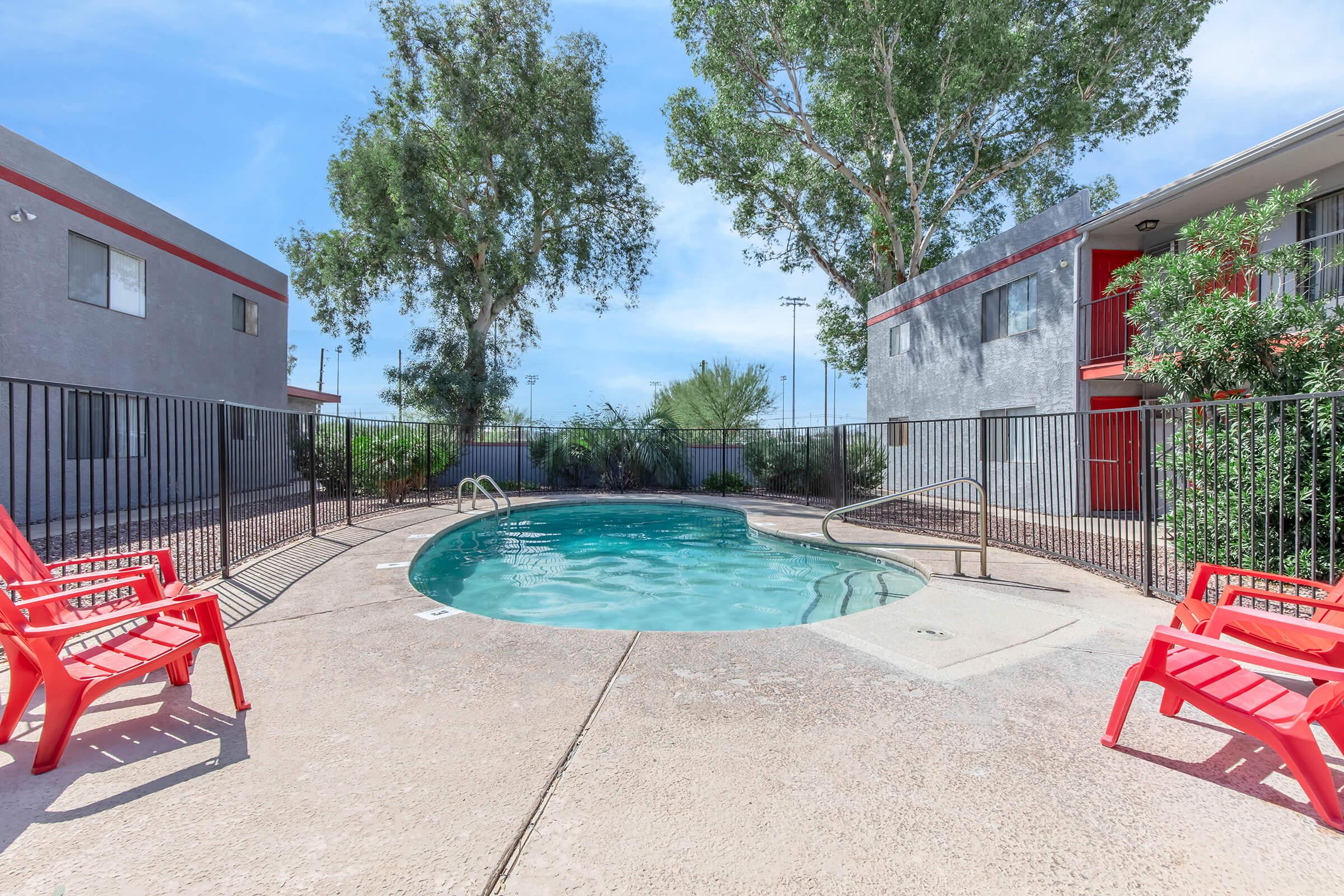 A small, oval swimming pool surrounded by a wrought-iron fence, with bright red lounge chairs placed nearby. The pool area is framed by landscaped greenery and is set against a backdrop of gray buildings with red accents. Clear blue skies and a few trees enhance the inviting atmosphere.